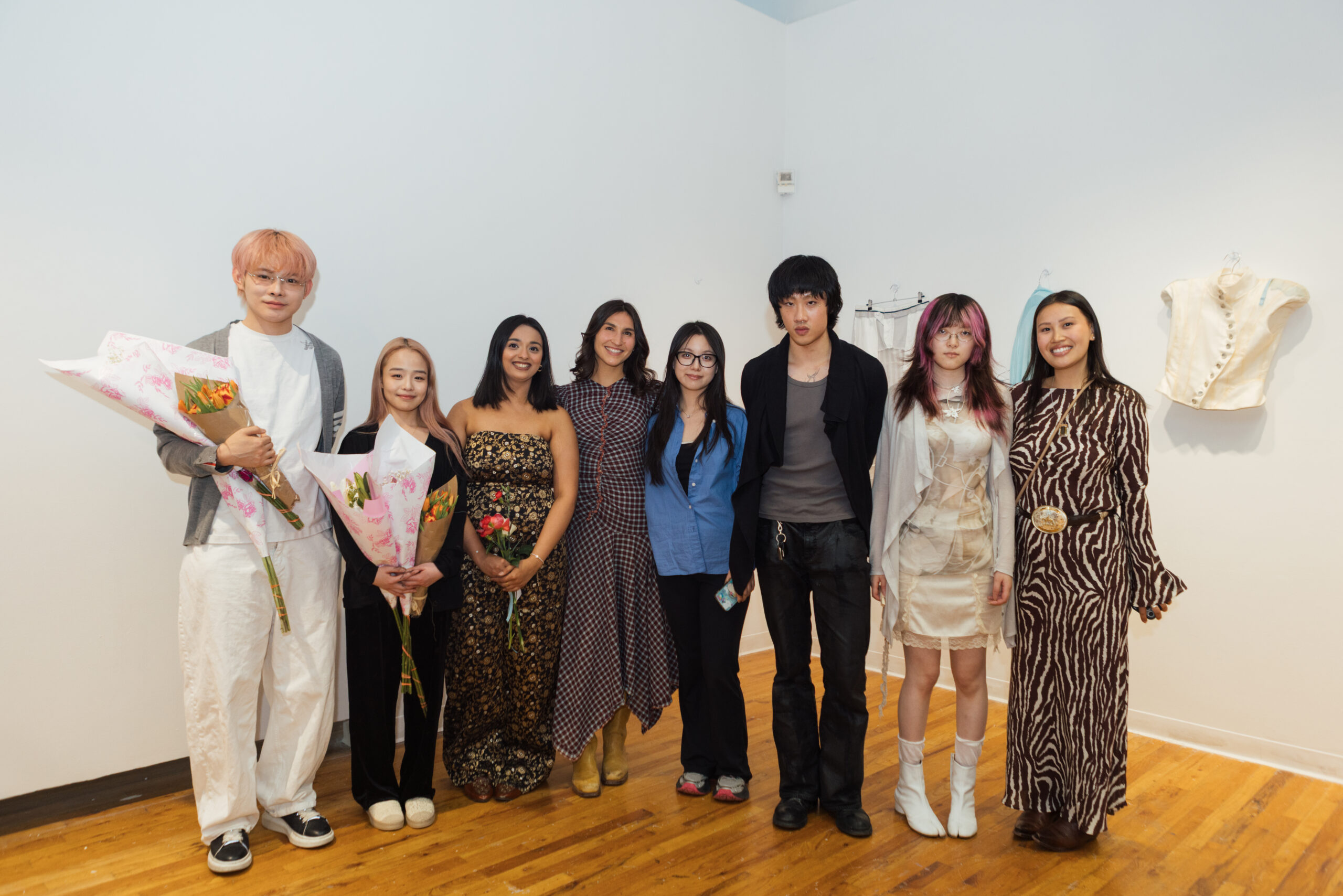 A group of seven young adults stands together in a gallery setting, smiling. They are dressed in various stylish outfits, with some holding bouquets of flowers. The background features a white wall with hanging clothing items. The floor is wooden, and the overall atmosphere appears cheerful and celebratory.