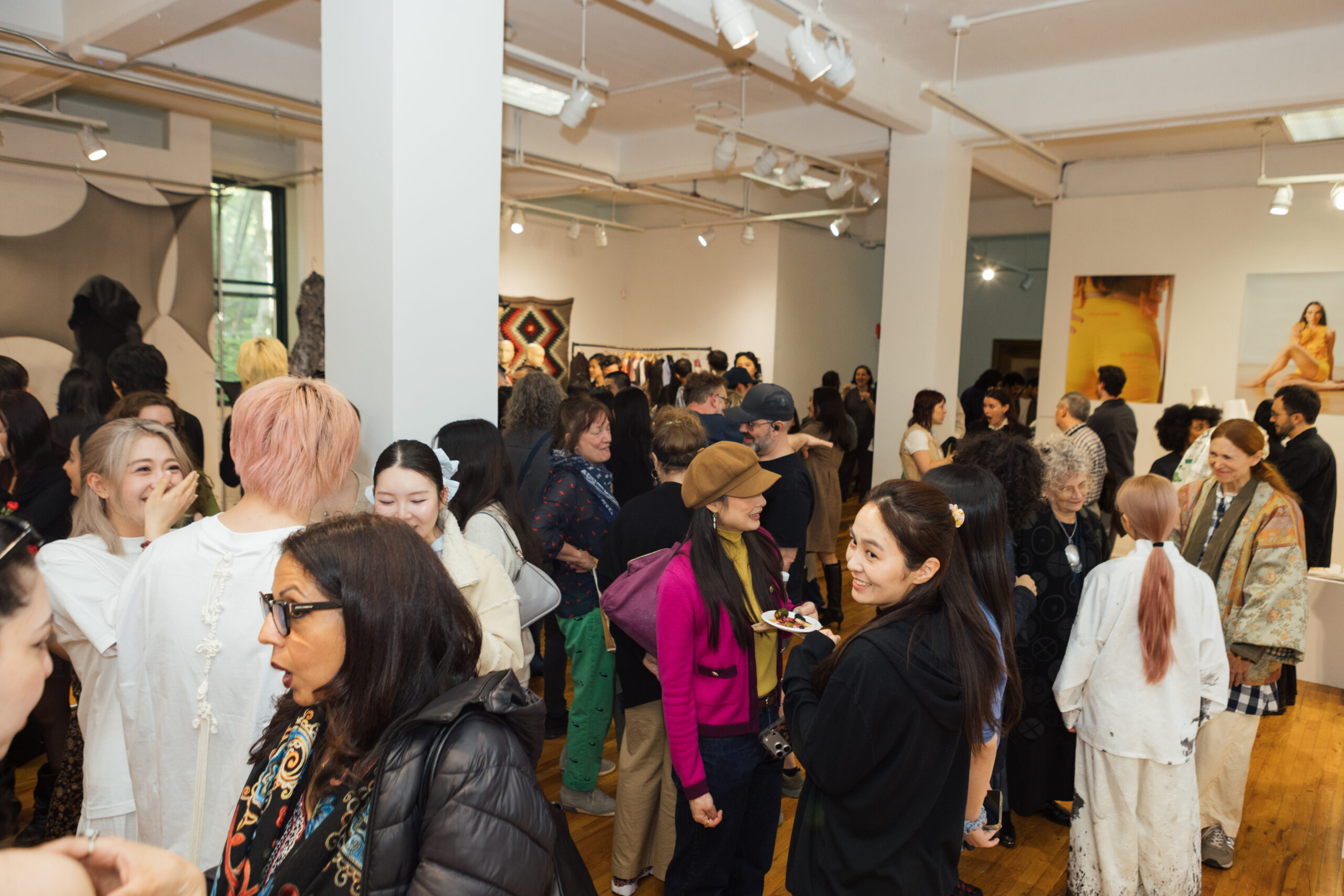 A lively art gallery event featuring a diverse group of attendees engaged in conversation. Individuals of various ages and styles are mingling, with some smiling and laughing. In the background, artwork and installations are visible, along with a few tables where refreshments may be served. The space has bright lighting and wooden flooring, contributing to a warm atmosphere.