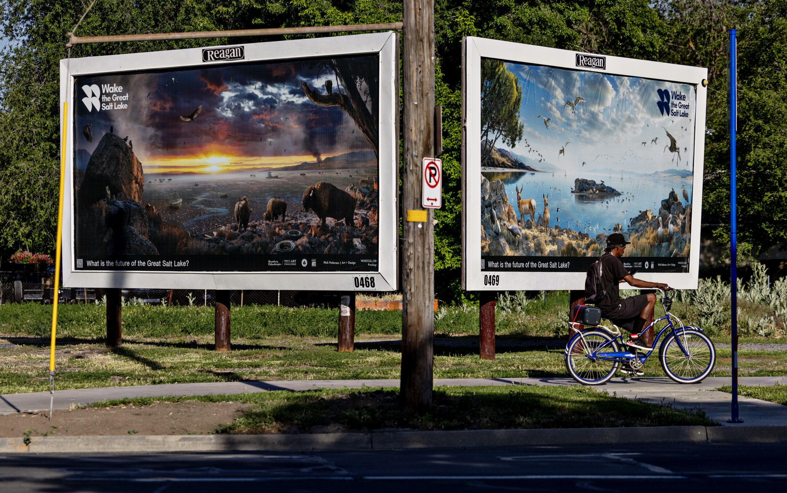 Two large billboards on a street display artwork related to the Great Salt Lake. The left billboard features a dramatic sunset over a landscape with wildlife, while the right shows a serene scene with water and animals. A person rides a blue bicycle on the sidewalk nearby. Green foliage and grass surround the area.