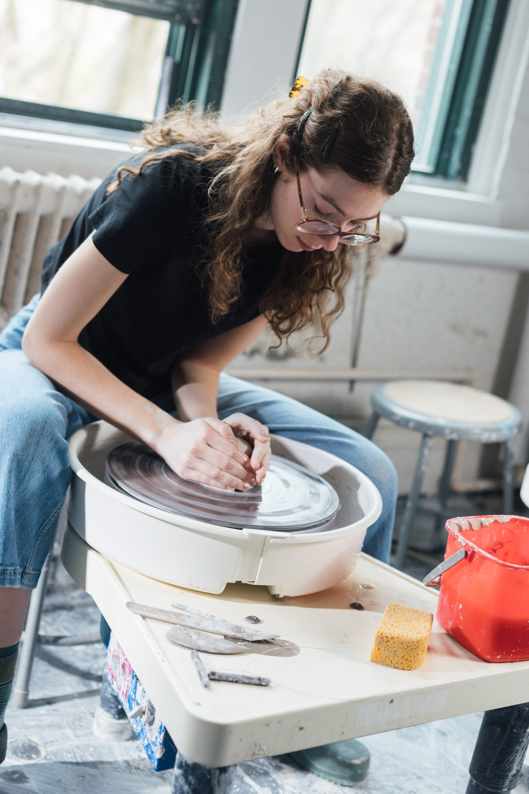 A person with curly hair is focused on shaping clay on a pottery wheel in a well-lit studio. They are wearing a black t-shirt and blue jeans. Nearby, a red bucket and clay tools are placed on the table, along with a yellow sponge. Large windows allow natural light to fill the room, highlighting the creative workspace.