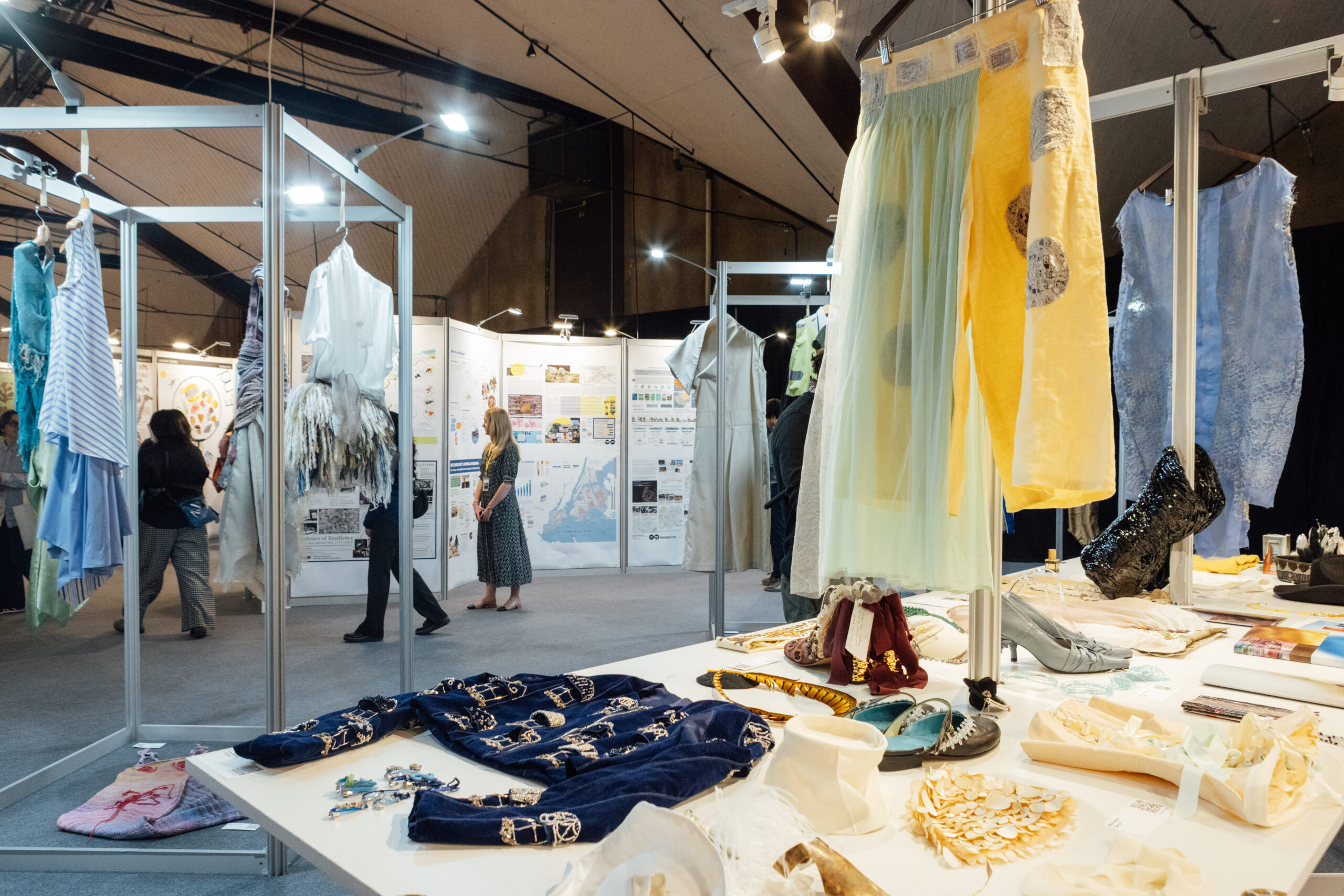 A fashion exhibition space featuring various clothing items displayed on racks and tables. In the foreground, blue velvet garments with embellishments and a variety of shoes are arranged on a table. In the background, attendees view clothing items and informational panels. The overall atmosphere is well-lit and vibrant, showcasing creative designs and accessories.