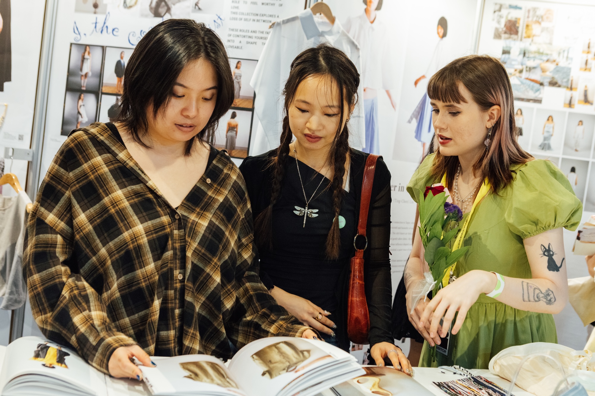 Three individuals are engaged in a discussion while examining a fashion book at an indoor event. The first person on the left is wearing a black and brown plaid shirt, the middle person wears a black top with a dragonfly pendant, and the third person on the right is dressed in a green puff-sleeved dress, holding a small bouquet of flowers. Behind them, there are posters and hanging garments displayed.