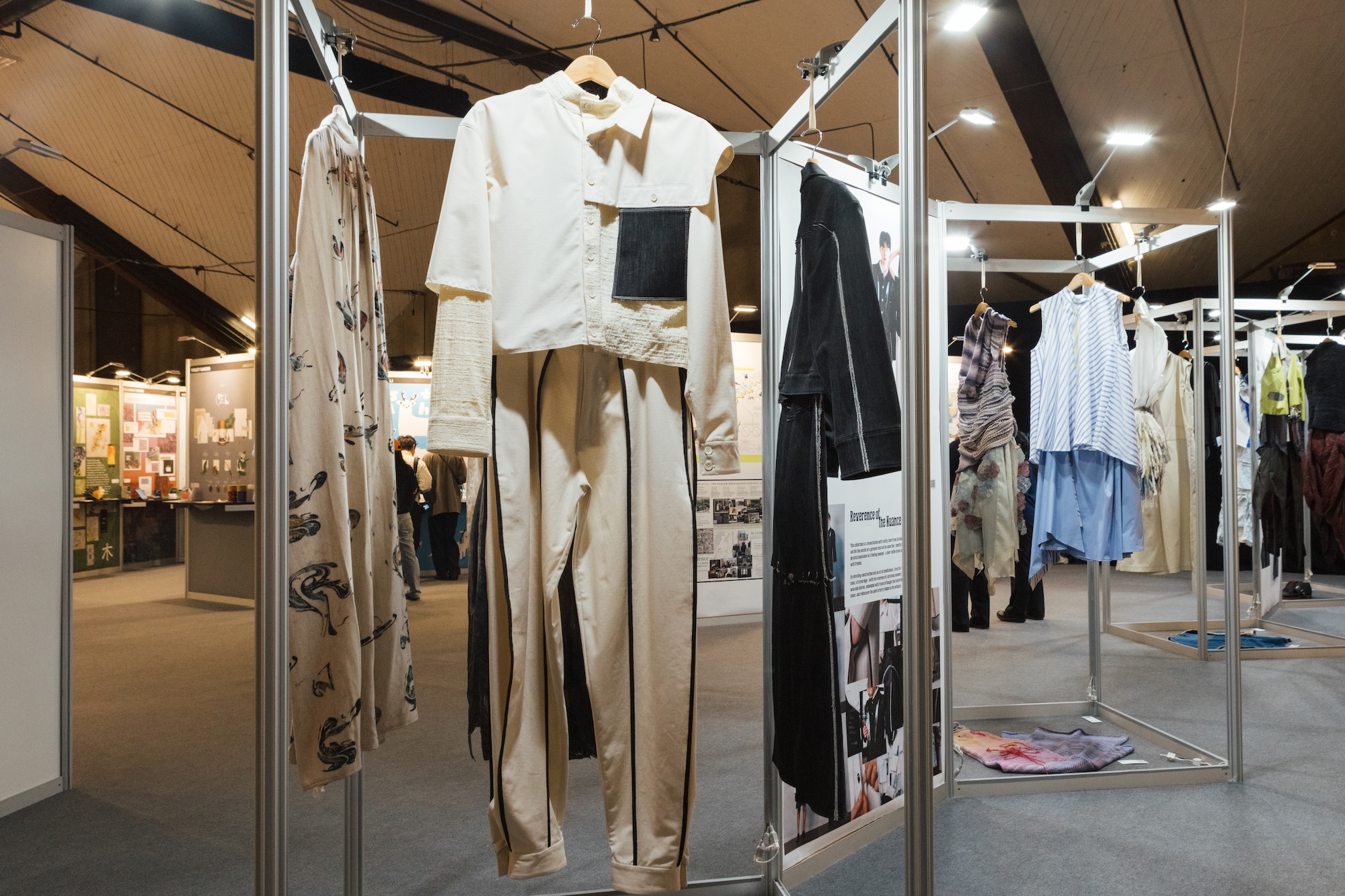 A fashion exhibition featuring various clothing displays on hangers, including a cream outfit with a cropped top and wide-legged pants, a black jacket, and a blue-striped shirt. The space is well-lit, with panels showcasing additional designs and illustrations in the background. Attendees can be seen interacting in the exhibition area.