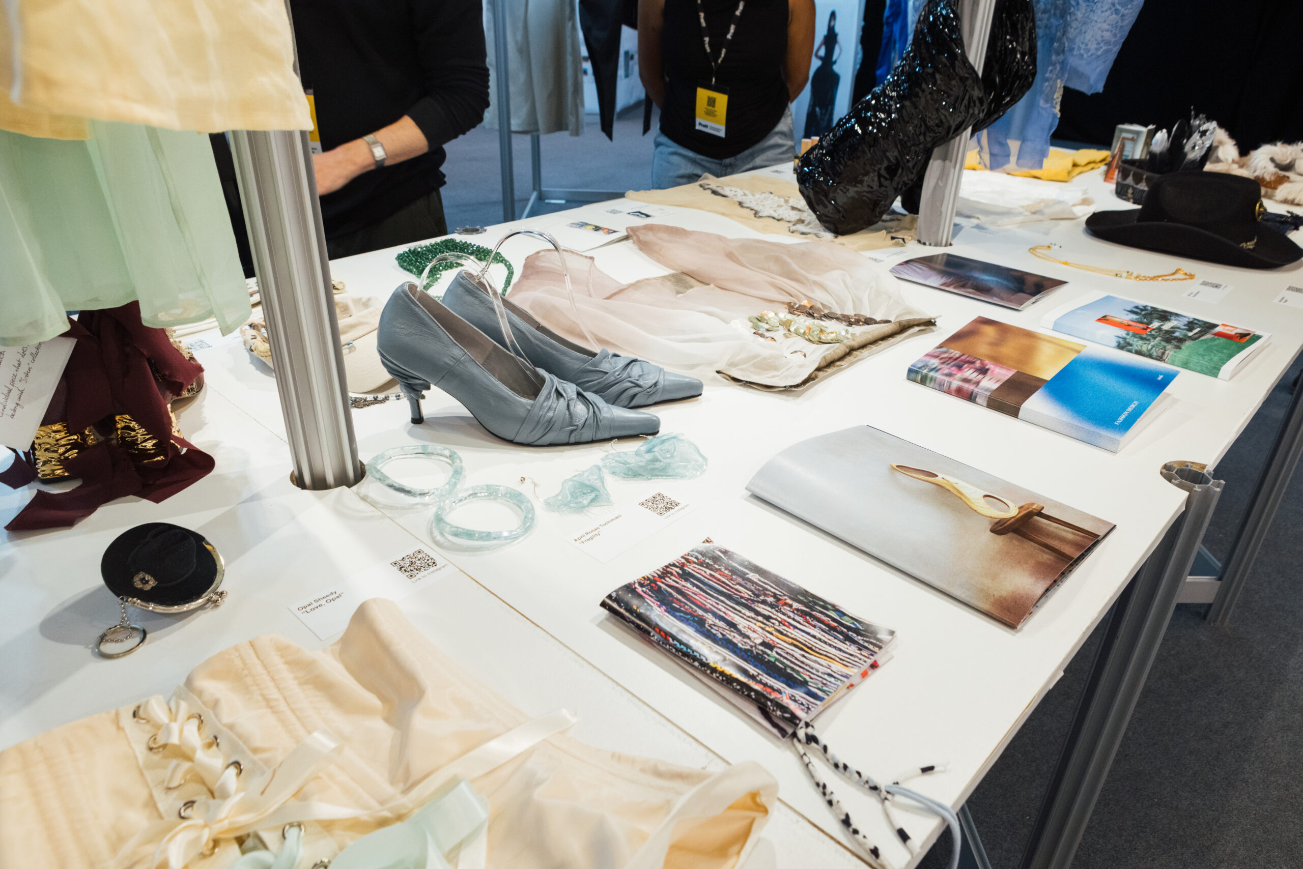 A display table features various fashion items. On the table are a pair of gray heels, several pieces of jewelry, including clear bangles, and a collection of fabric swatches. There are also magazines showcasing fashion designs and accessories. The background shows people interacting with the display, with some clothing items hanging nearby.