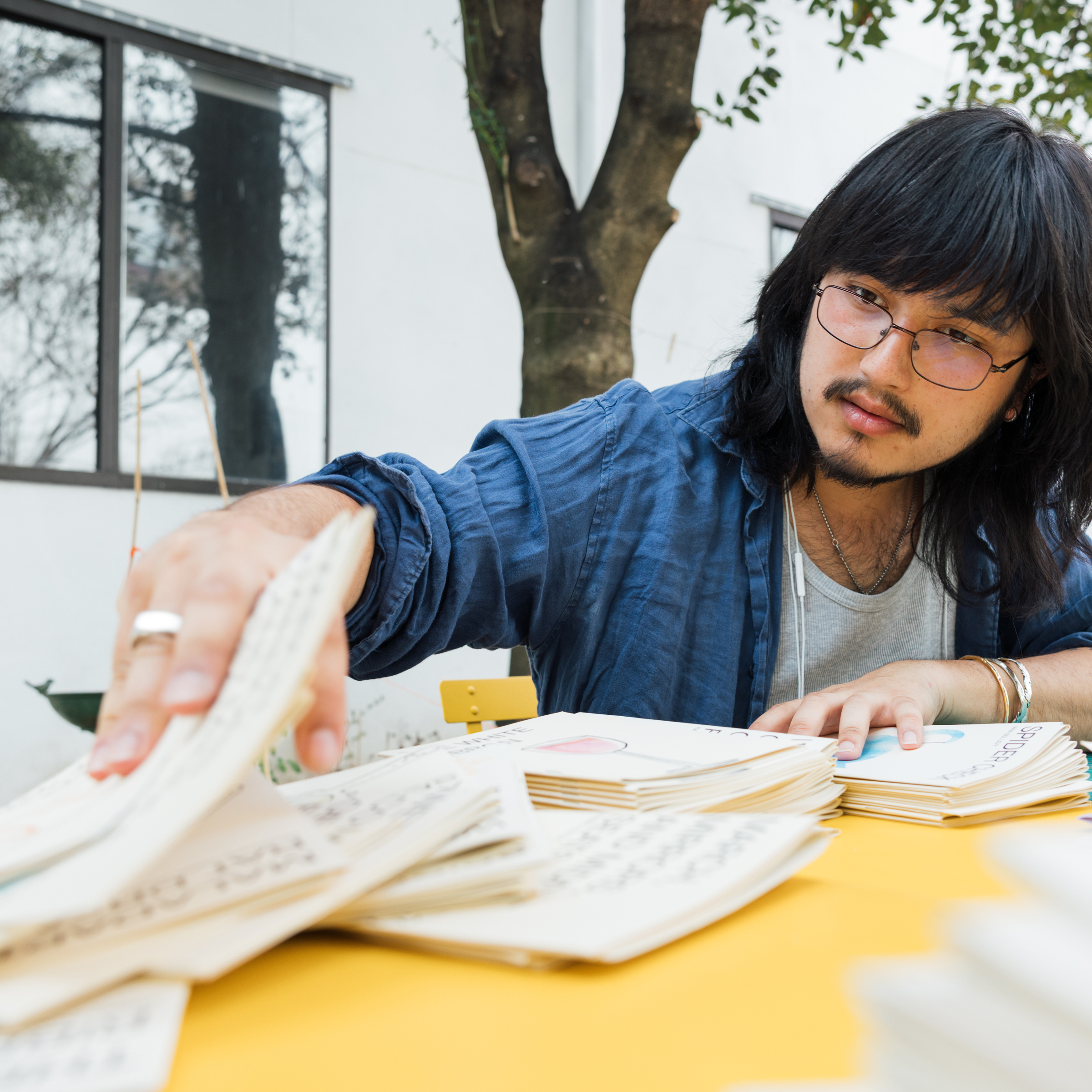 A person with long black hair and glasses is focused on sorting through a stack of papers on a yellow table outdoors. The background includes a tree and a building with large windows. The individual is wearing a blue shirt over a gray top, with earphones around their neck.