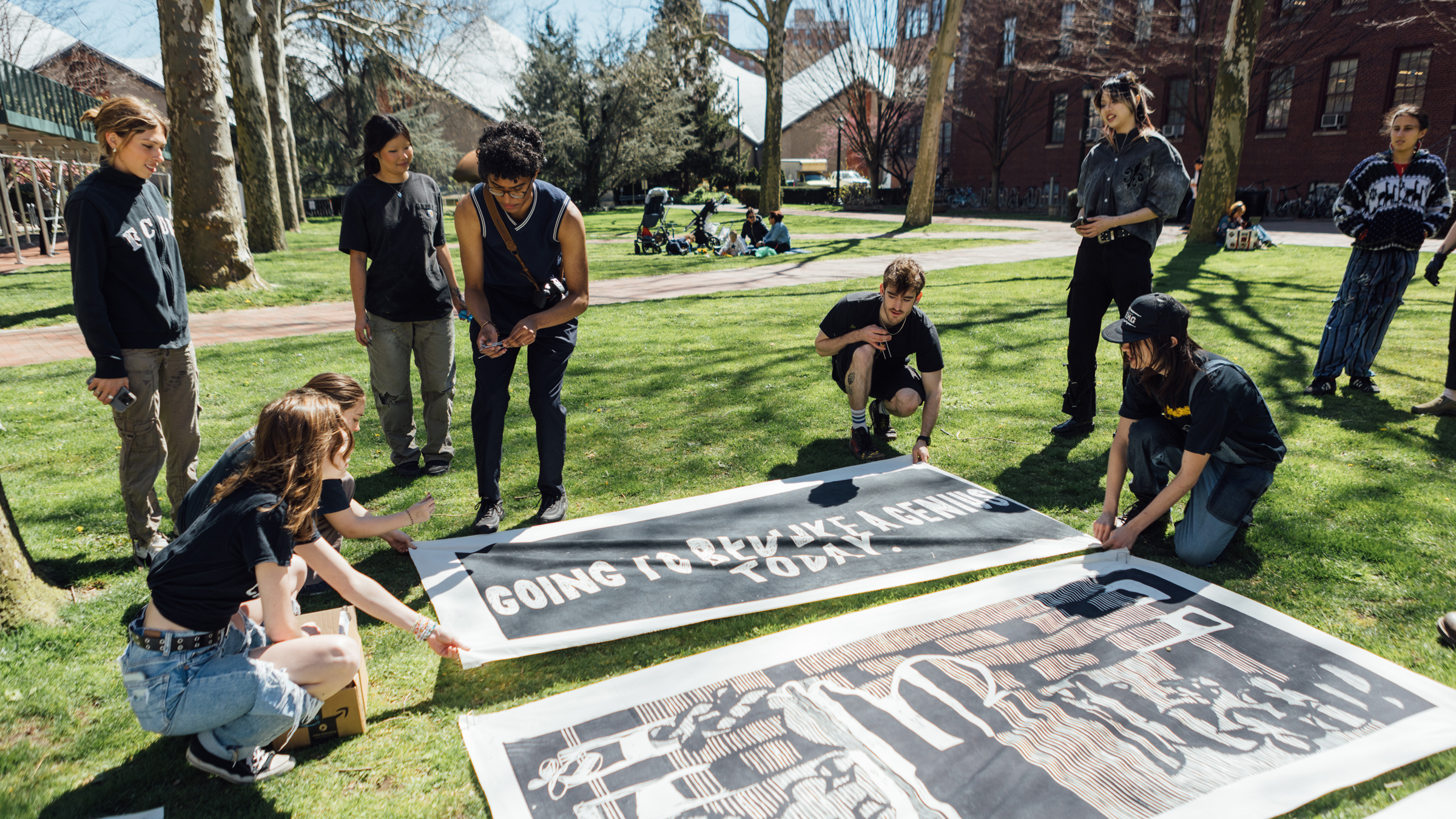 A group of young people prepares large banners on a grassy area under clear blue skies. Some individuals are kneeling or crouching as they position the banners, which feature bold text and illustrations. Other members of the group stand nearby, monitoring the setup. In the background, additional people are leisurely engaged in activities. The atmosphere appears collaborative and energetic.