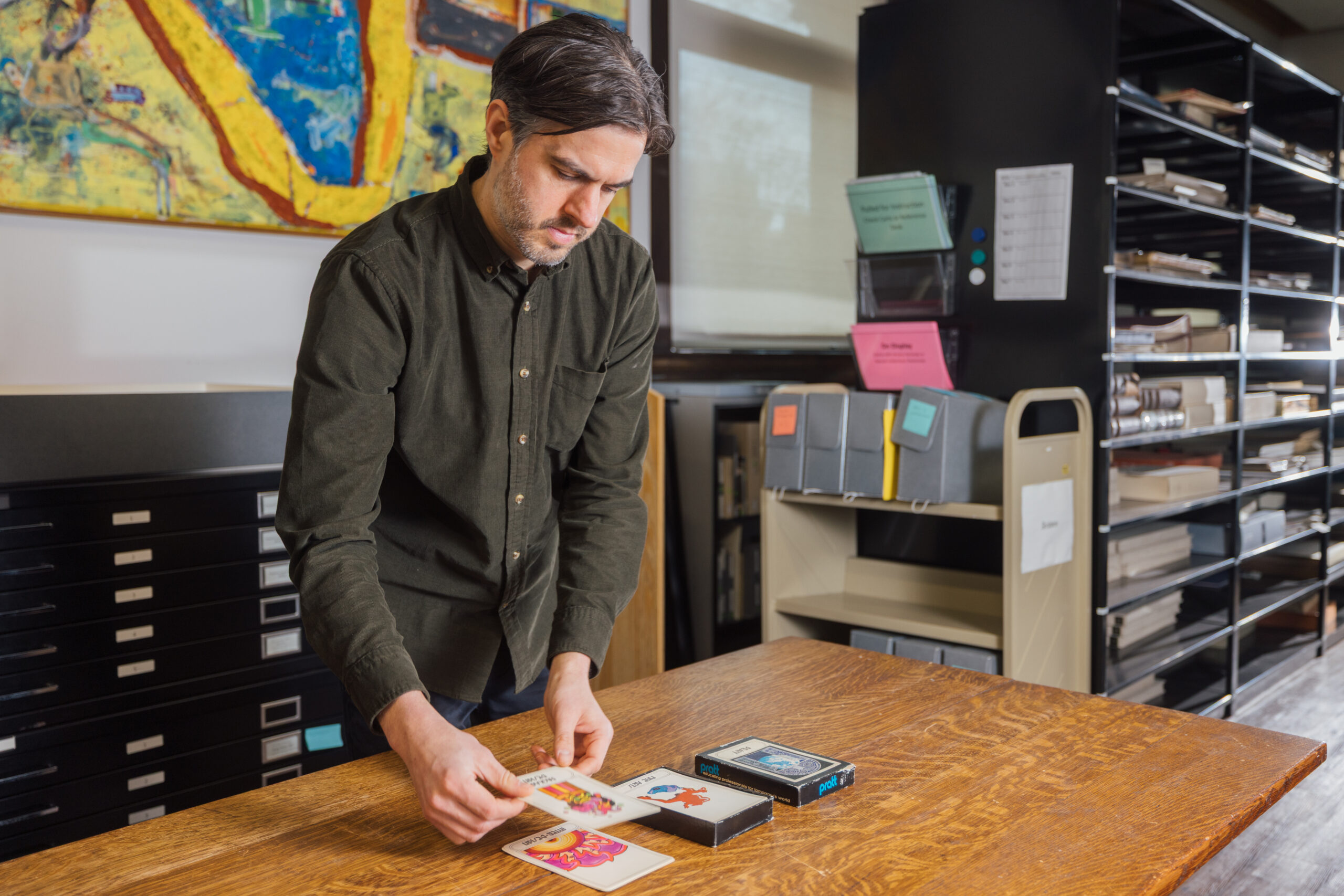 A man with dark hair and a beard is organizing several colorful postcards on a wooden table in a library or archival setting. He is wearing a dark green button-up shirt and appears focused on his task. In the background, there are shelves filled with various folders and boxes, along with an abstract painting on the wall. The environment suggests an organized space for document or artifact handling.