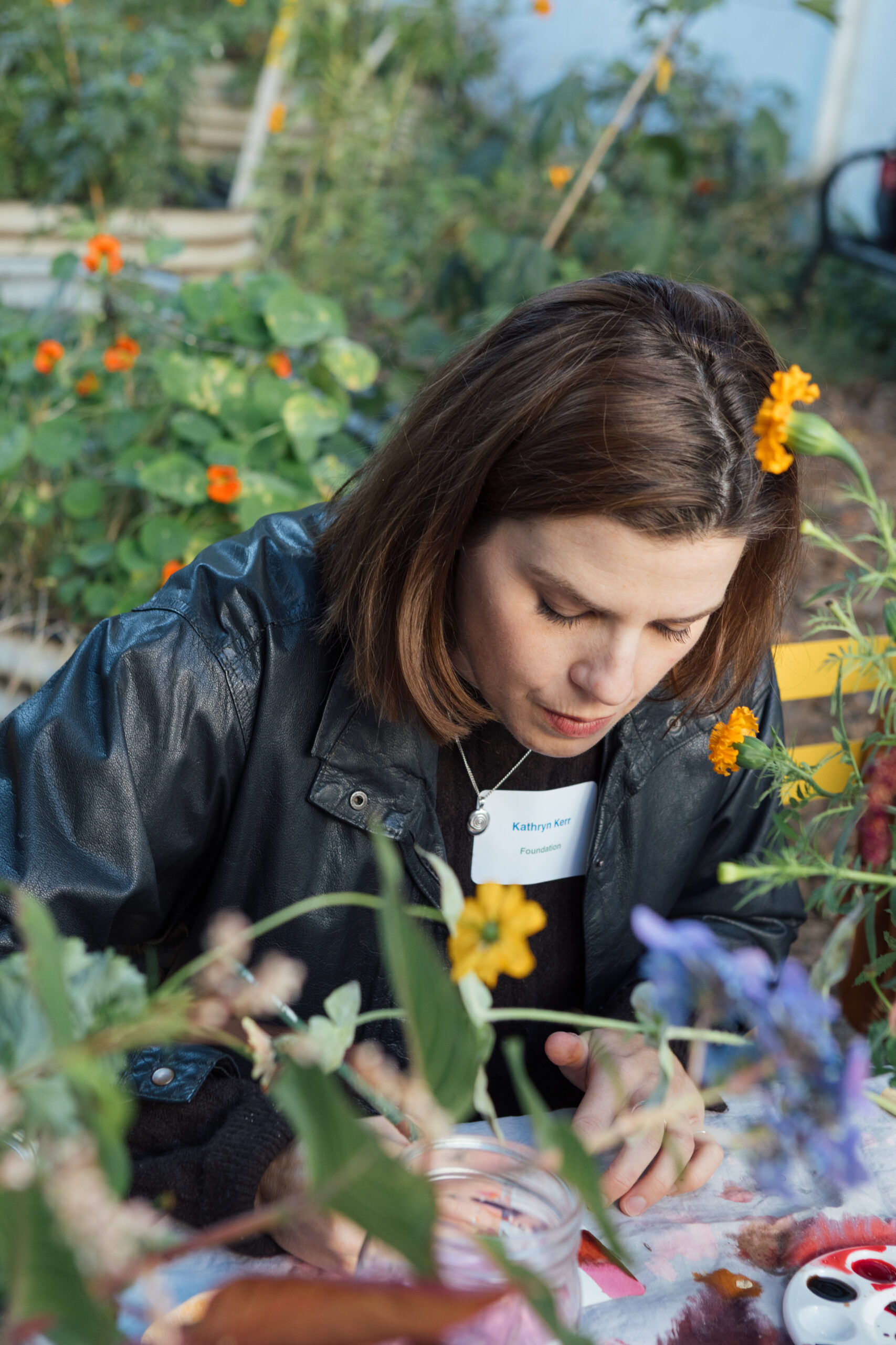 A woman with short brown hair is seated at a table surrounded by colorful flowers. She is focused on a task, lightly leaning forward with a paintbrush in her hand. She's wearing a black leather jacket and a name tag with the name "Kathryn Kerr" and the word "Foundation" visible. The setting is outdoors, likely in a garden or greenhouse, with vibrant greenery and blooming flowers in the background.