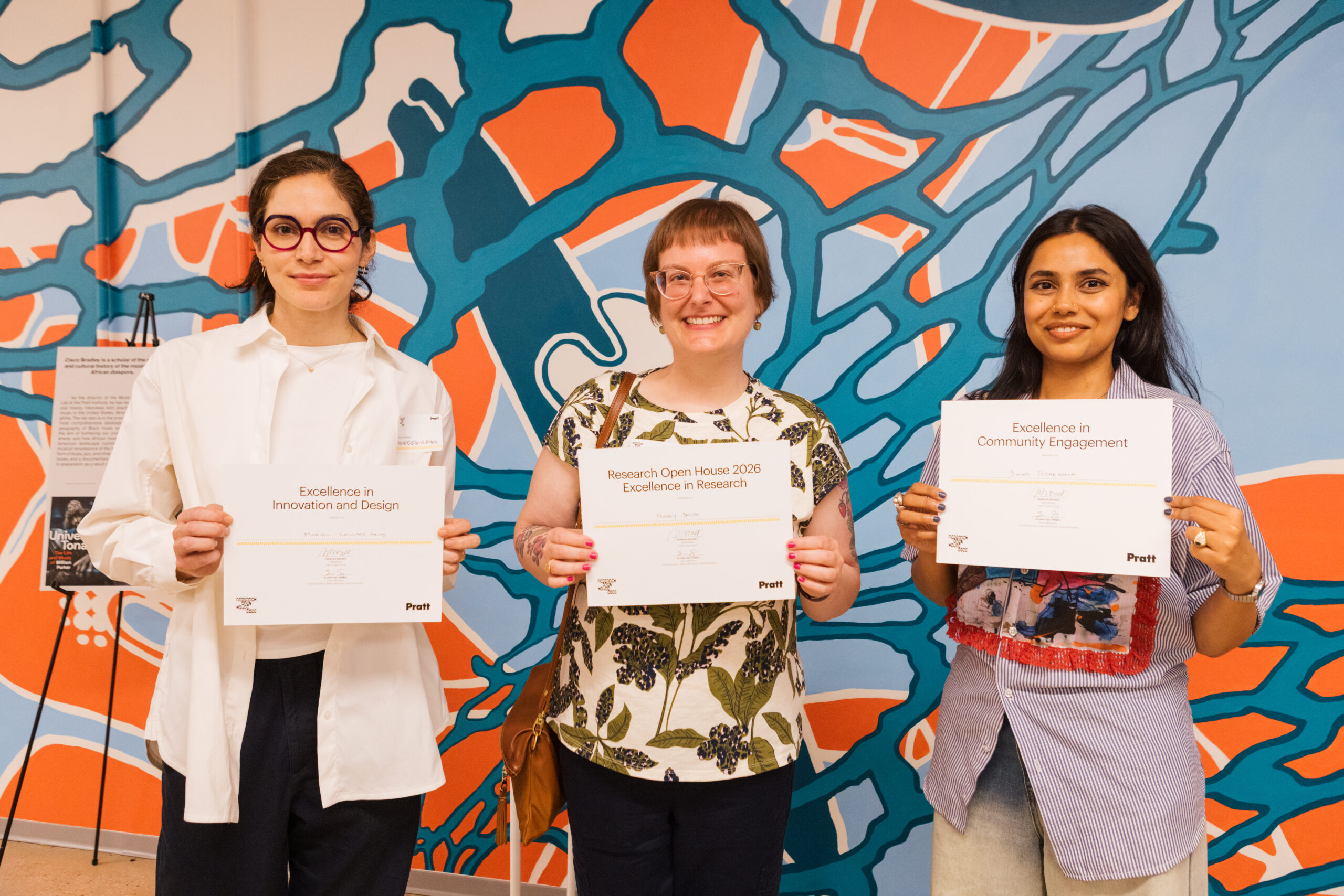 Three individuals are standing together, each holding a certificate related to their achievements. The woman on the left wears glasses and a white lab coat, holding a certificate for "Excellence in Innovation and Design." The woman in the middle has short hair, is smiling, and holds a certificate for "Excellence in Research." The woman on the right, dressed in a striped shirt, is holding a certificate for "Excellence in Community Engagement." The background features a colorful abstract mural.