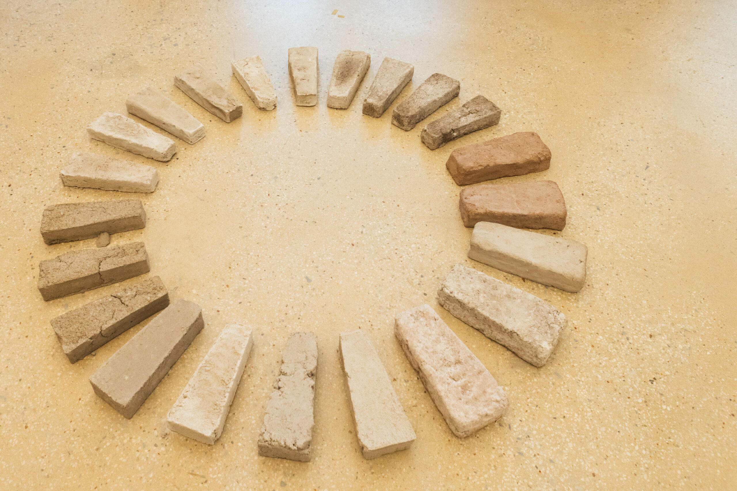 A circular arrangement of rectangular bricks in various shades, ranging from light beige to dark brown, is laid out on a smooth, pale yellow floor surface. The bricks are placed closely together, forming a continuous loop, and display different textures and colors.