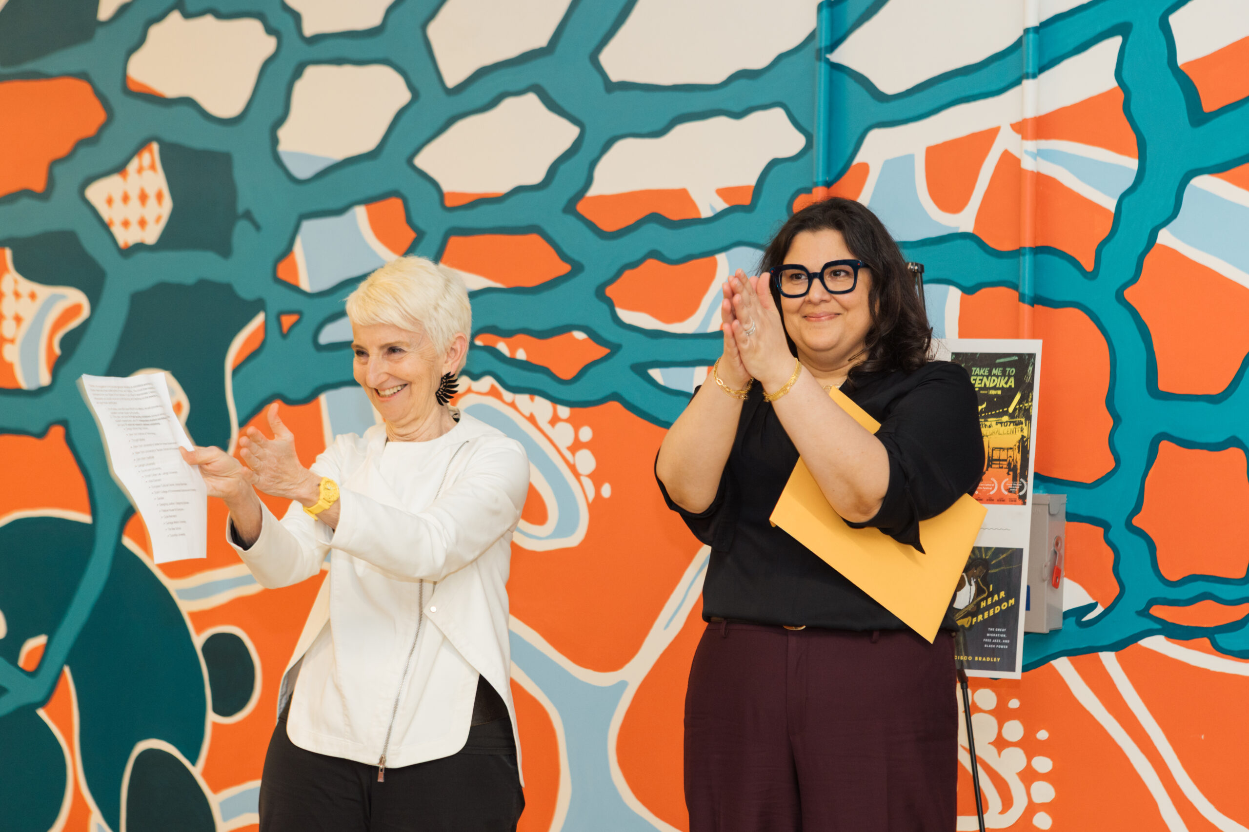 Two women are standing in front of a colorful mural. The woman on the left is smiling and holding a sheet of paper, while the other woman on the right is clapping and holding a yellow folder. The background features a vibrant design with teal and orange patterns.