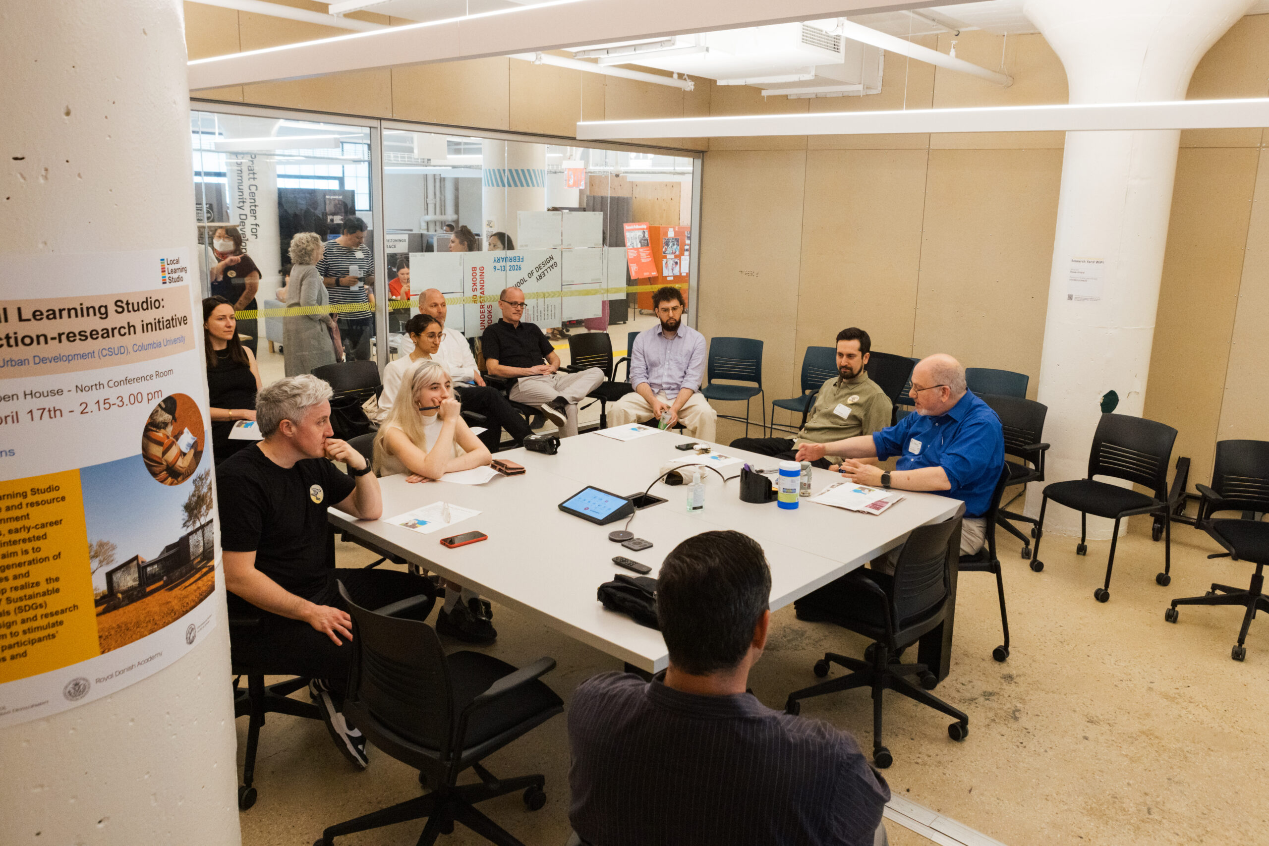 A group of people is seated around a large conference table in a modern, well-lit room. The gathering appears focused on discussion, with participants taking notes and listening attentively. A poster is visible on a nearby pillar, detailing an event titled "Spatial Learning Studio: Action-research initiative." Several individuals are visible, including a person with short hair and a black shirt, a woman with light hair, and a man in a blue shirt speaking.