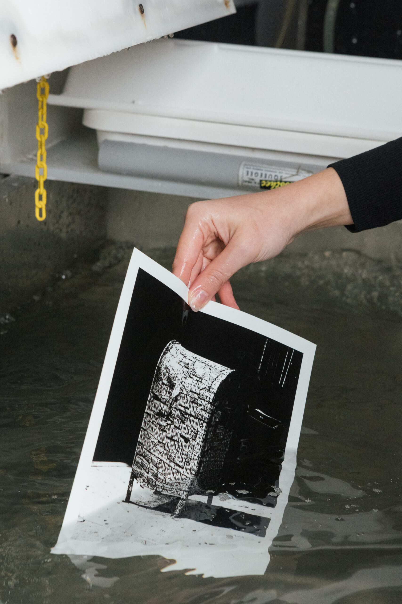 A hand is holding a black and white photograph partially submerged in water. The image depicts a textured object, possibly a basket or container, with text visible on its surface. The background features water ripples and reflections, indicating a wet environment.