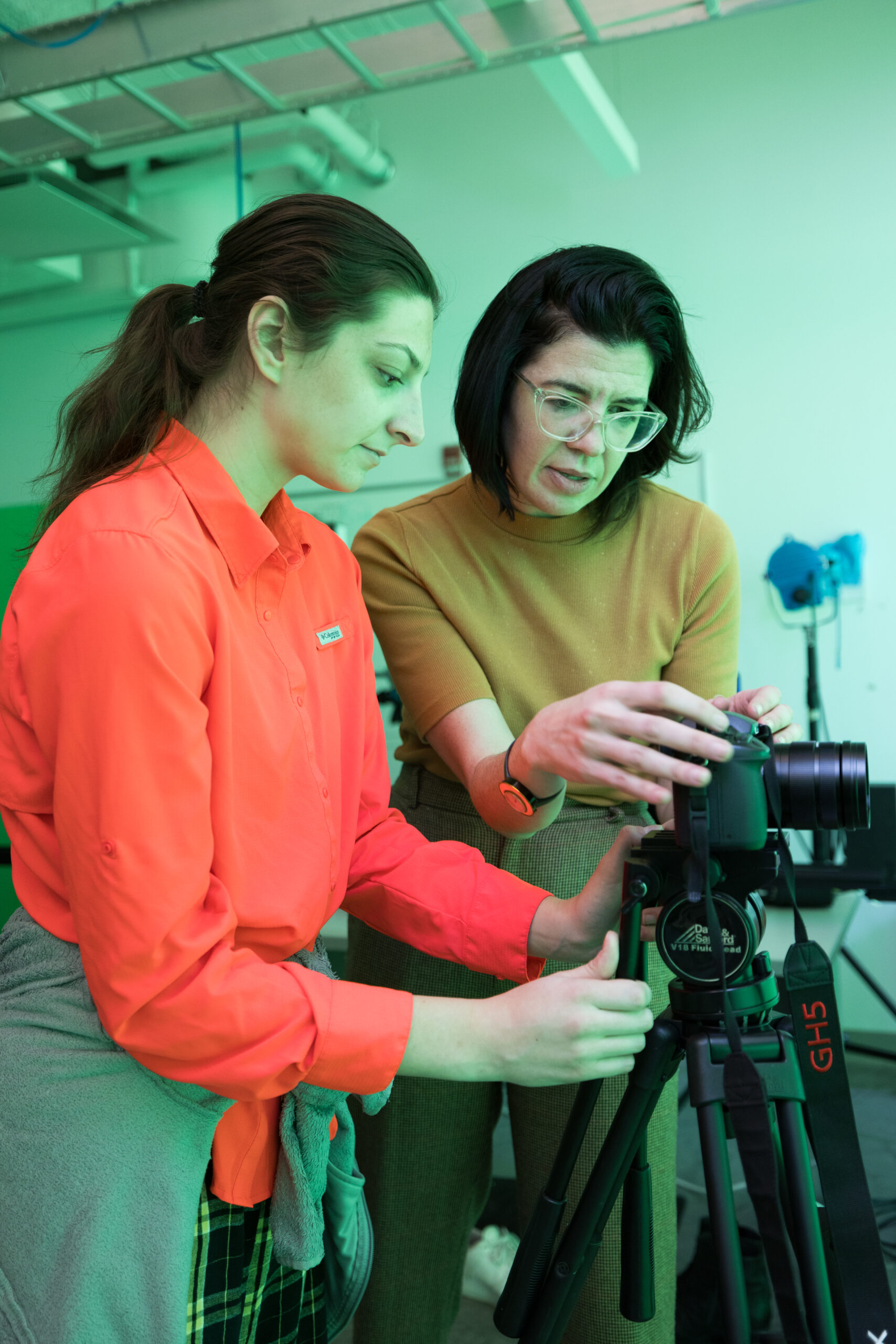 Two women are closely examining a camera set on a tripod in a brightly lit studio. One woman, wearing an orange shirt, gestures while holding the camera, and the other, in a brown top with glasses, observes intently. The background features green hues, with some studio equipment visible.
