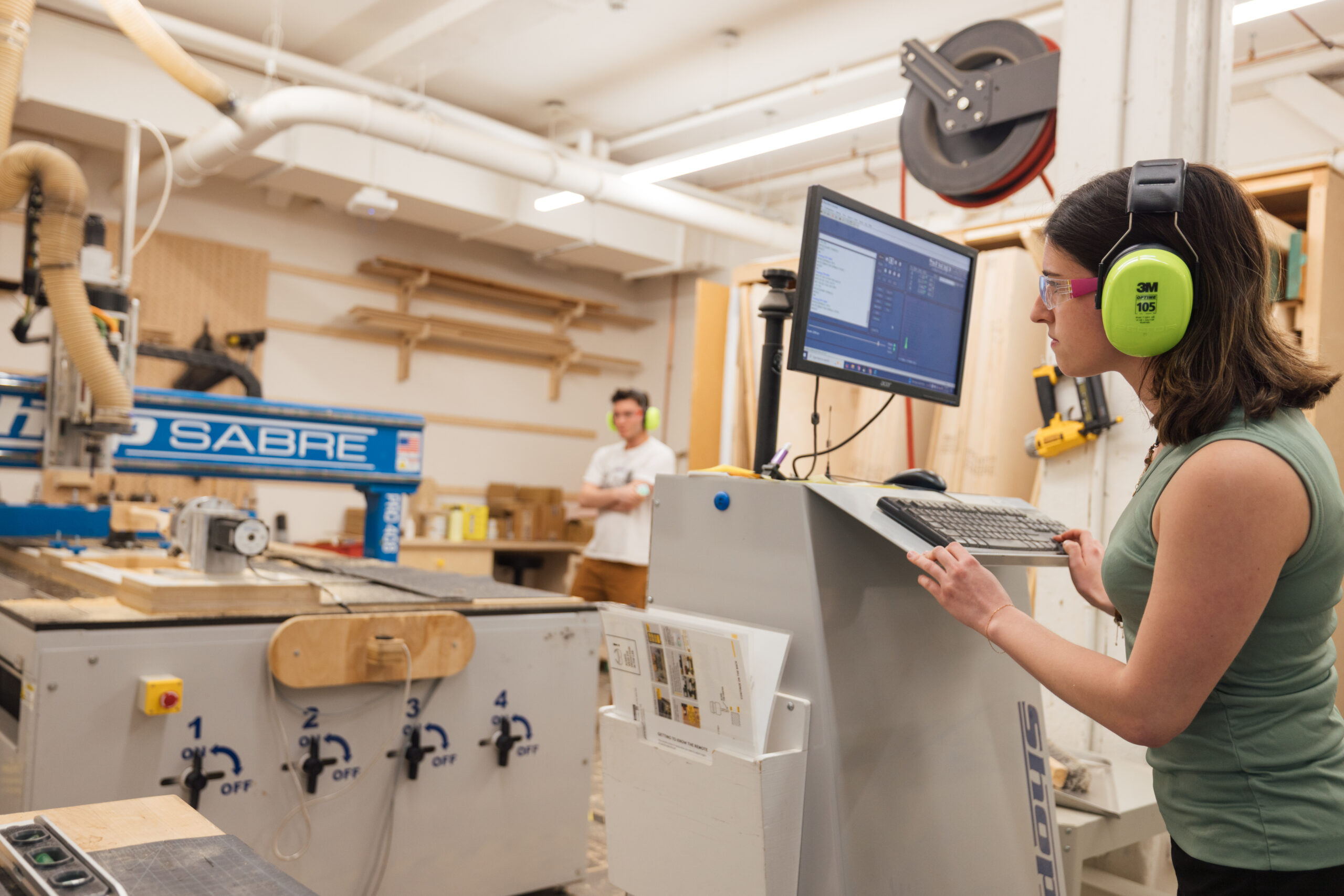 A woman wearing safety glasses and bright green ear protection operates a computer terminal in a woodworking shop. She is focused on the monitor displaying software controls. In the background, a man stands with his arms crossed, observing. A large CNC machine labeled "SABRE" is set up nearby, and various woodworking materials are visible on shelves and tables.