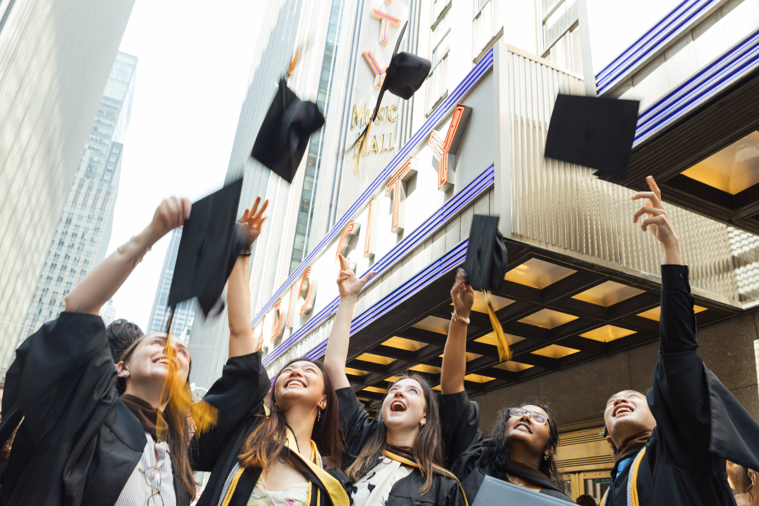 A group of graduates in black caps and gowns joyfully throw their caps into the air outside a theater, celebrating their graduation. The scene is bright, with tall buildings and the marquee of the theater visible in the background. The graduates have wide smiles, conveying a sense of achievement and happiness.