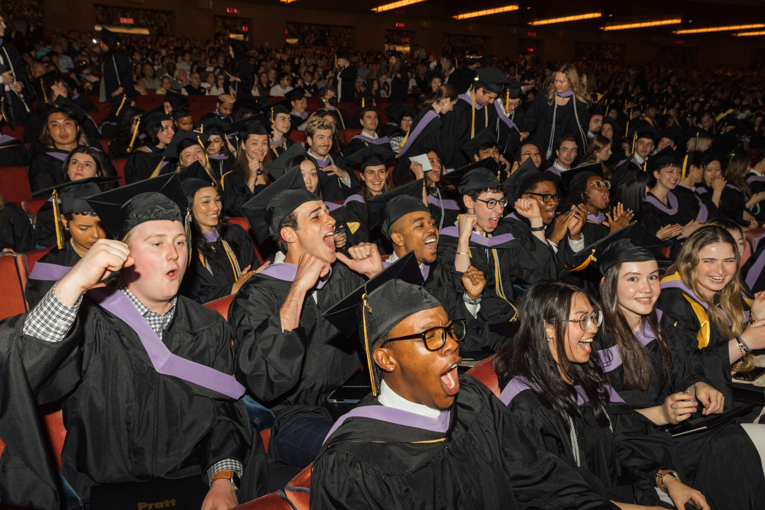 A large group of graduates in black gowns and caps is cheering and celebrating in a theater during a graduation ceremony. Many are smiling and clapping, showing excitement. The audience is packed, with some individuals visibly expressing joy and enthusiasm.