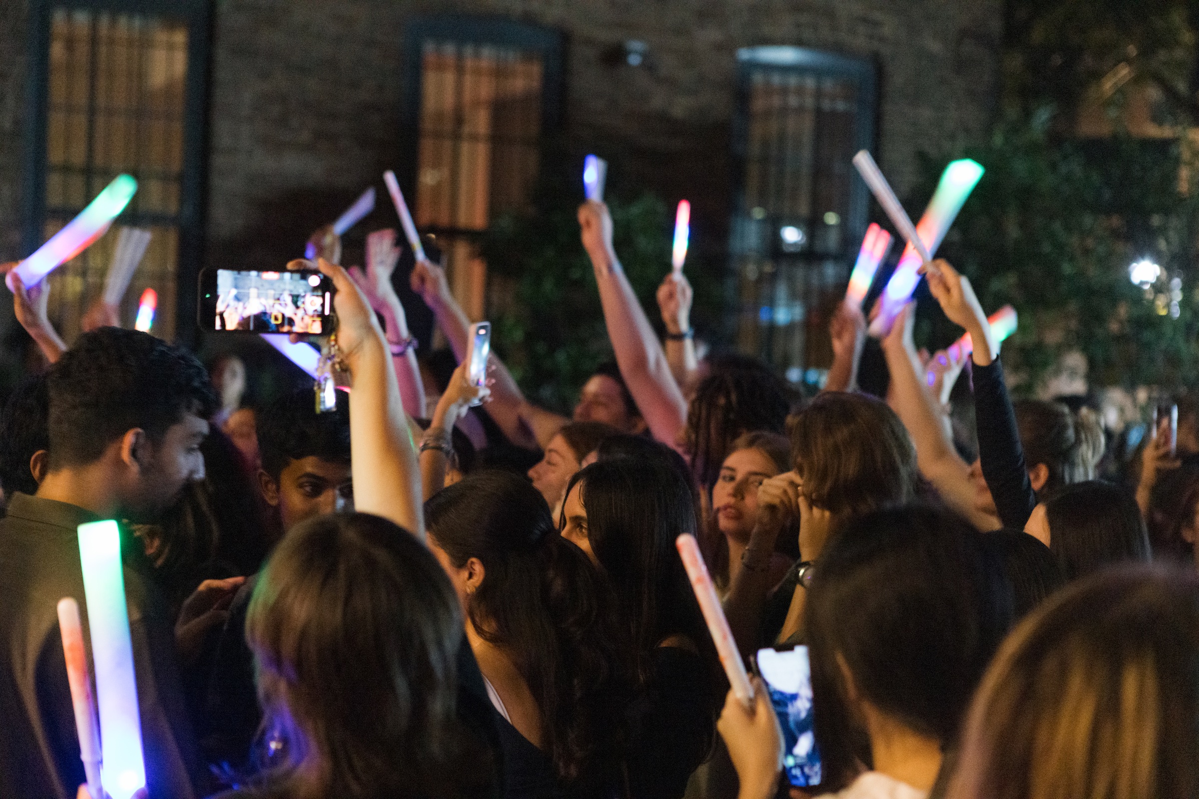 A vibrant crowd of young people is gathered outdoors, illuminated by colorful glow sticks. Many individuals are holding their phones up to capture the moment, while others engage with one another. The atmosphere is lively, with people smiling and enjoying the event, surrounded by a backdrop of brick walls and soft lighting.