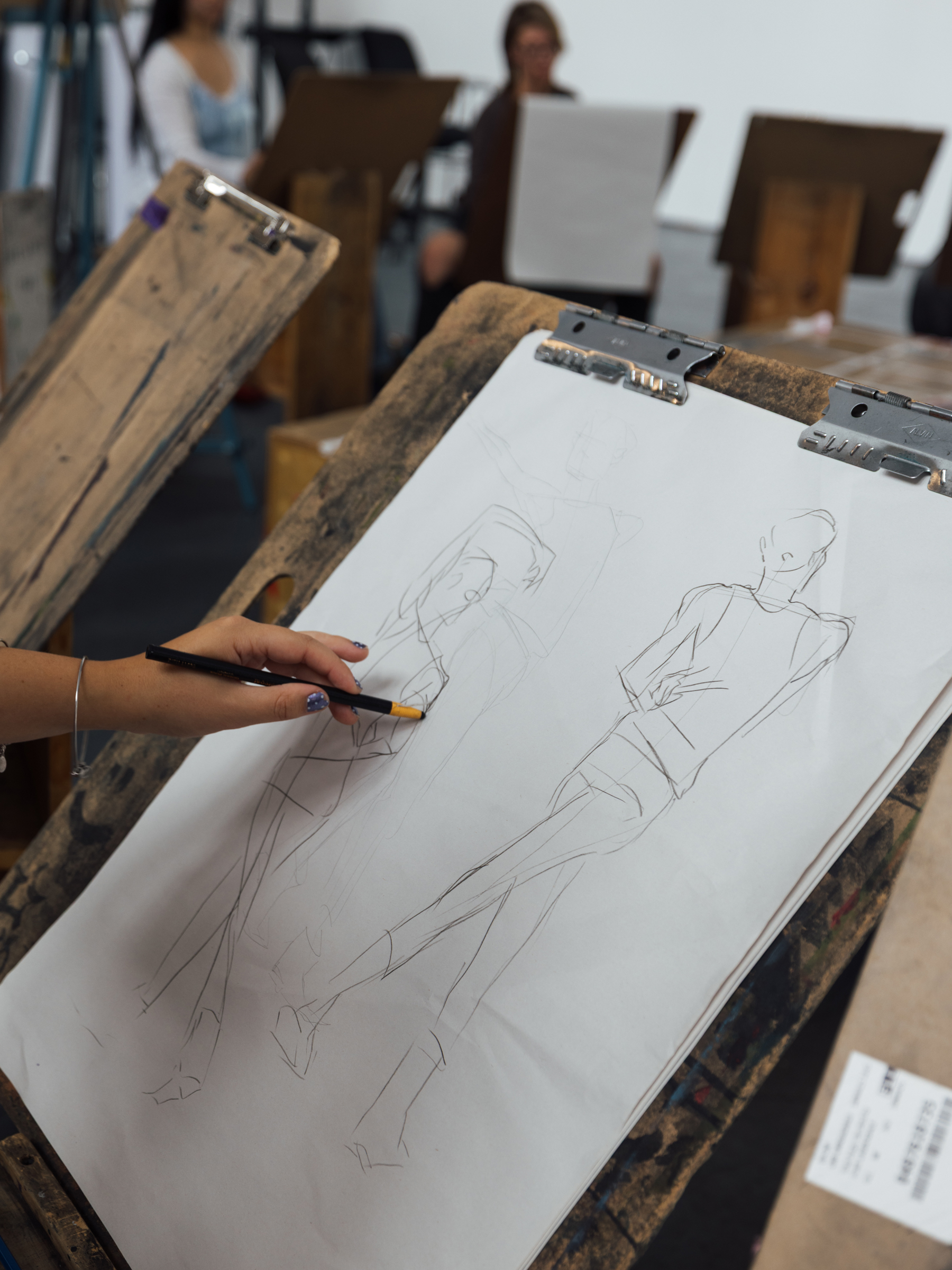 A partially completed drawing is displayed on an easel, showing rough sketches of human figures. A hand holding a pencil is seen in the foreground, actively working on the drawing. In the background, other artists are seated at similar easels, focused on their work in an art studio setting.