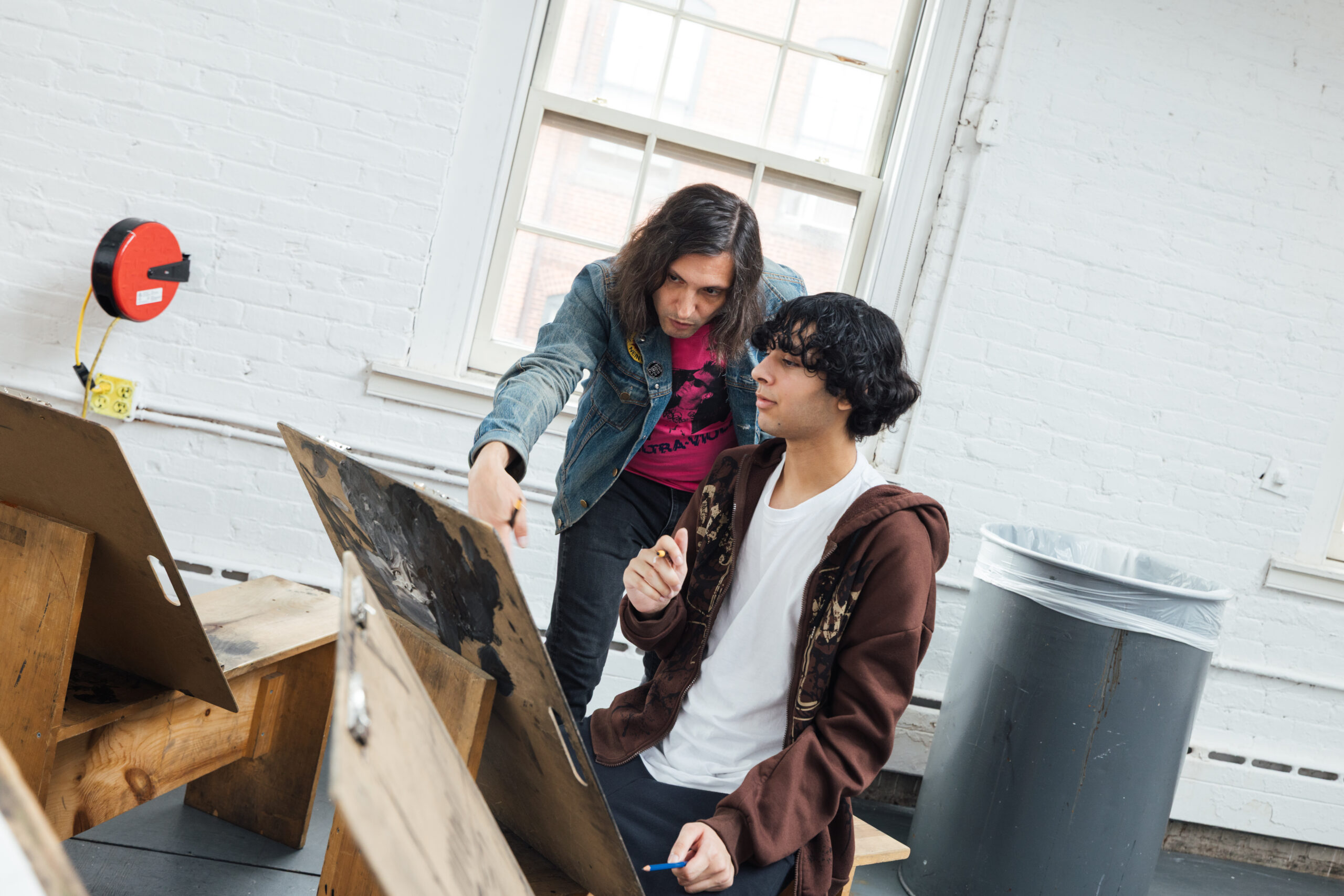 Two individuals are engaged in an art studio setting. One person, wearing a denim jacket and a pink shirt, is standing beside another seated individual, who is wearing a brown hoodie and a white shirt. The standing person appears to be giving guidance or feedback, pointing towards an easel or workstation in front of the seated individual, who is holding a pencil and looking attentively. In the background, there is a trash bin and large windows allowing natural light to enter the room.