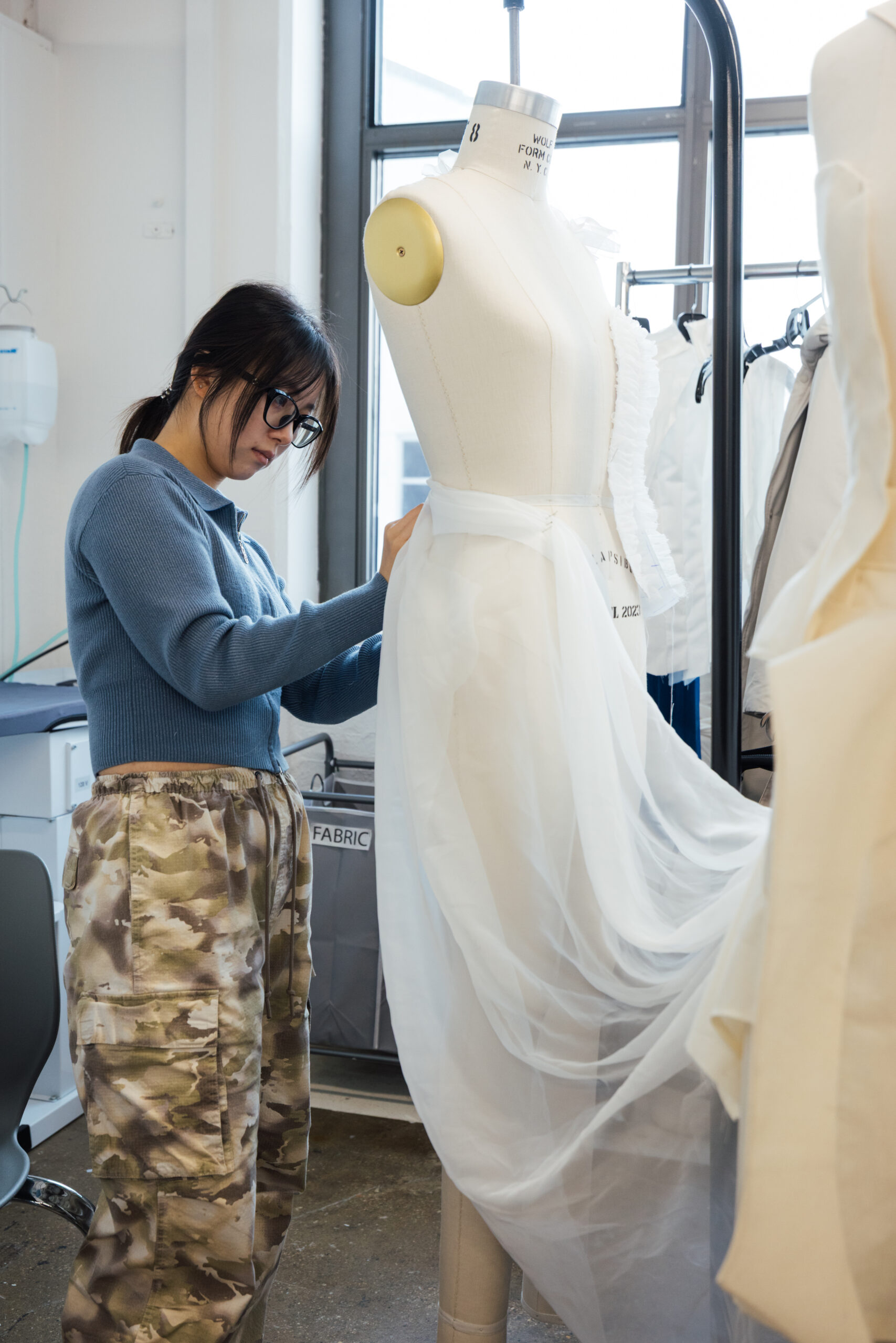 A person wearing a blue sweater and camouflage pants adjusts a white tulle skirt on a dress form in a fashion studio. Bright natural light filters through a window, highlighting the workspace, which includes fabric draped on nearby racks and a printer in the background.