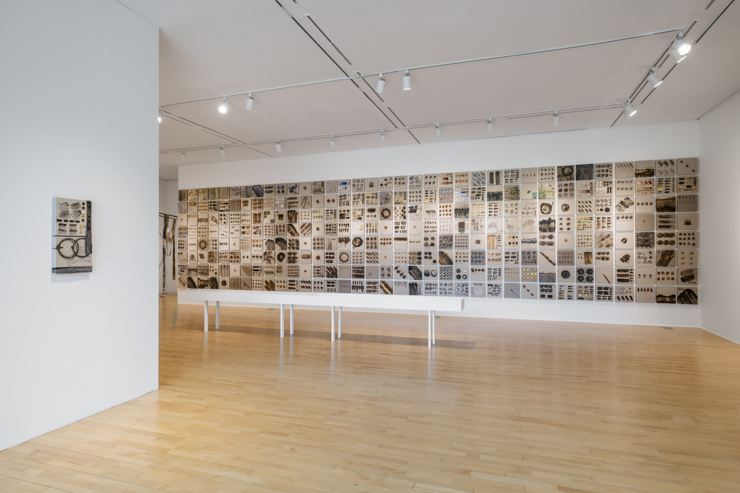 An art exhibition space featuring a large wall collage filled with numerous panels displaying various items, likely artifacts or illustrations. In the foreground, there is a small artwork mounted on the wall, and a long white bench is positioned in front of the wall, providing a space for viewers to sit and contemplate.