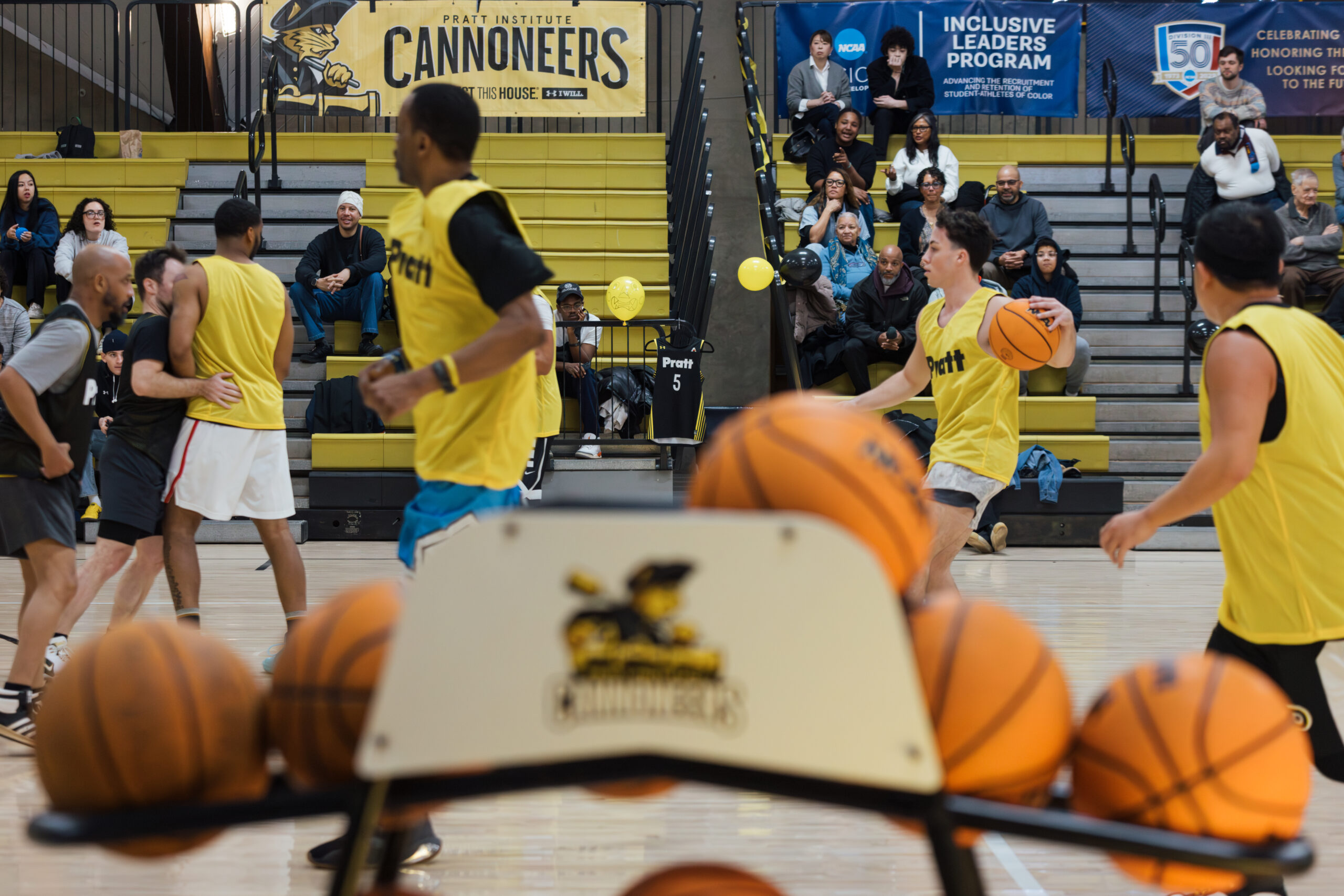 A basketball game is taking place in a gymnasium. Players wearing yellow jerseys are running down the court, while a line of basketballs is positioned in the foreground on a rack. In the background, several spectators are watching the practice from the bleachers. A banner for the Pratt Institute Cannoneers hangs on the wall.