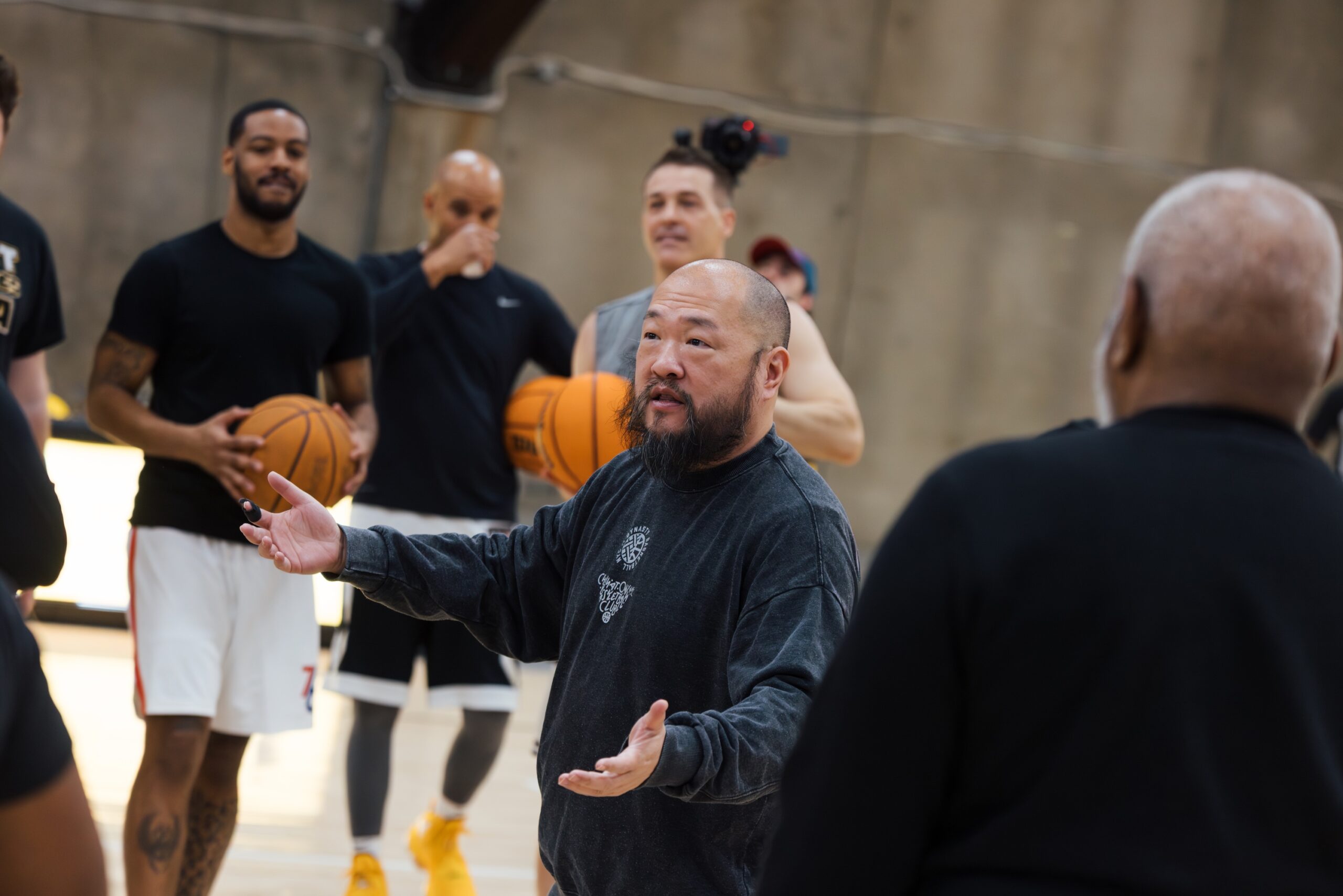 A group of people in a basketball gym, with one player, Bernard Chang, speaking and gesturing with his hands. He has a beard and is wearing a dark sweatshirt. In the background, several participants are holding basketballs.