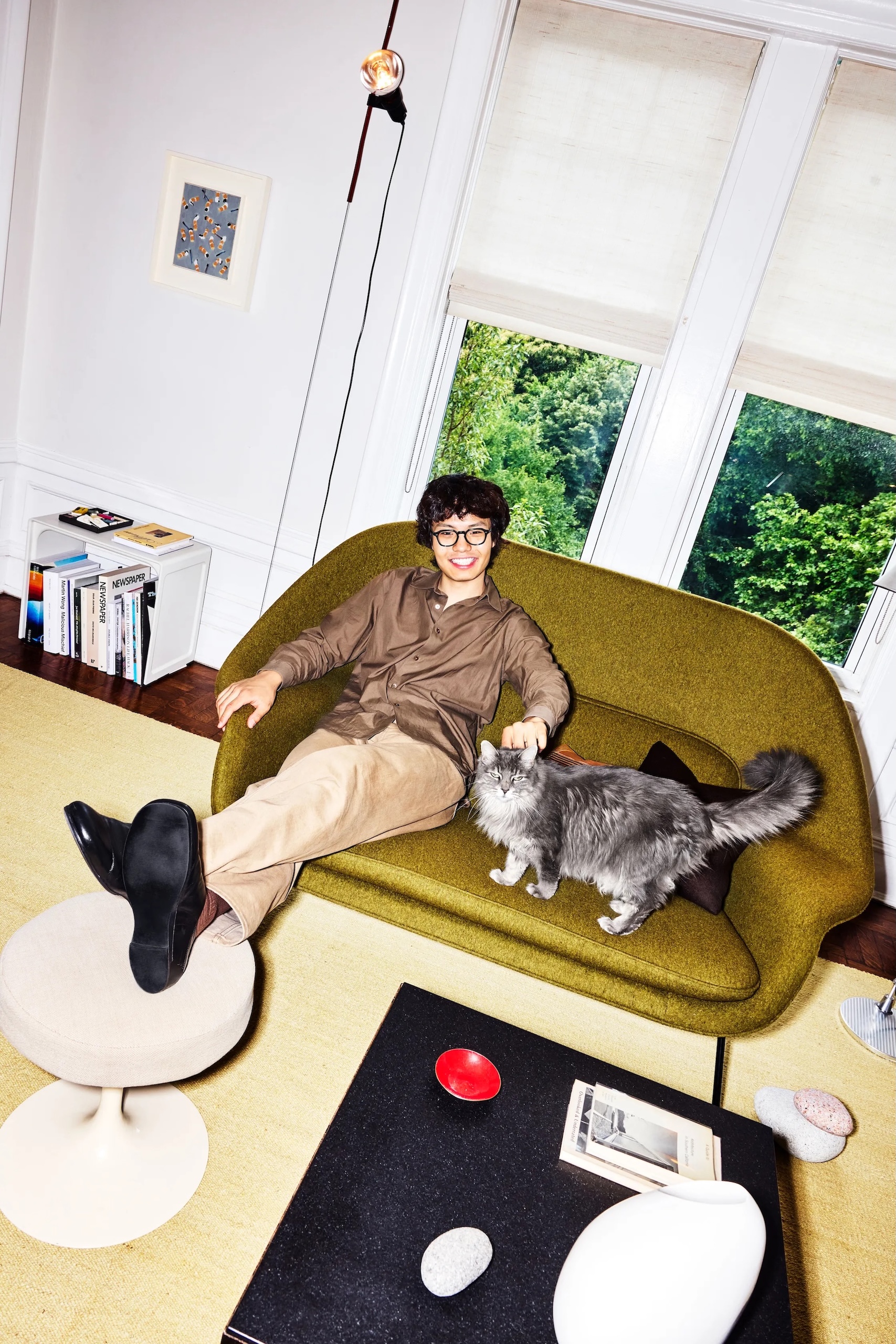 Jason Chen sits on an olive green sofa, smiling while petting a gray cat beside them. They wear a brown shirt and light-colored pants. In the background, there are large windows showing greenery outside. A small white table with a round top is in the foreground, holding decorative stones and a red bowl. A black coffee table is also visible, featuring a book and more stones.