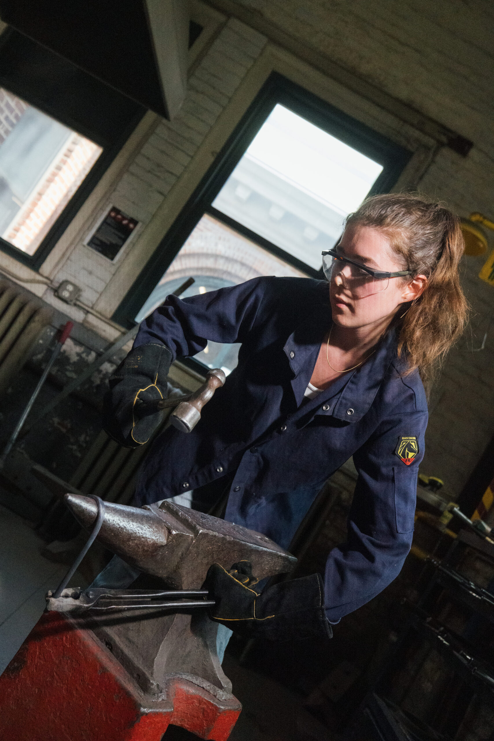 A person working at a blacksmith station, hammering a piece of metal against an anvil. They are wearing protective eyewear and gloves, and a navy blue work jacket. The background shows windows with brickwork, and various tools are visible in the workspace.