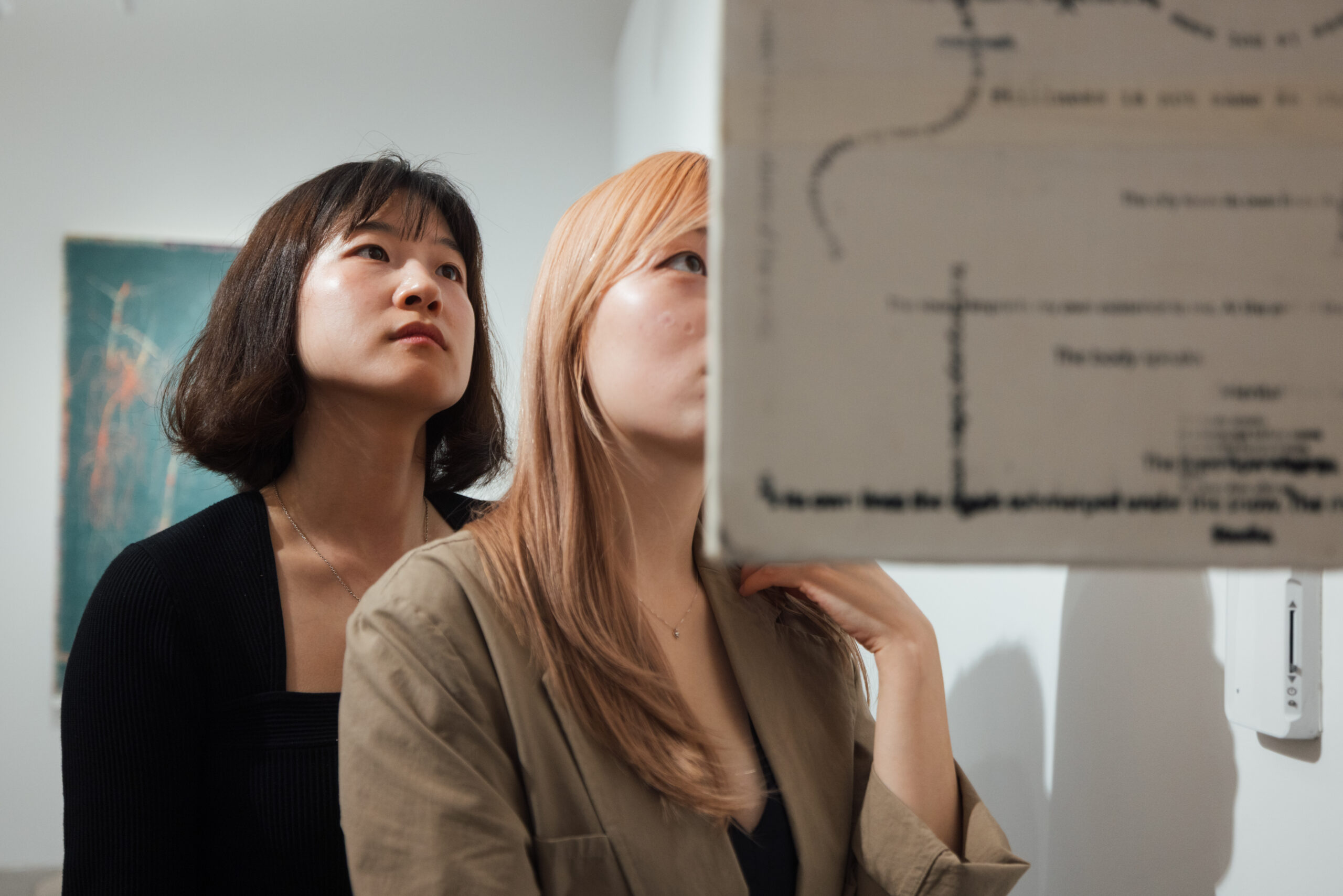 Two young women are looking upwards, engaged in viewing an exhibit in an art gallery. The woman in front has short, dark hair, while the one behind has long, light-colored hair. A partially visible display in front of them contains text and lines. The background includes an abstract painting. The atmosphere appears contemplative and focused.