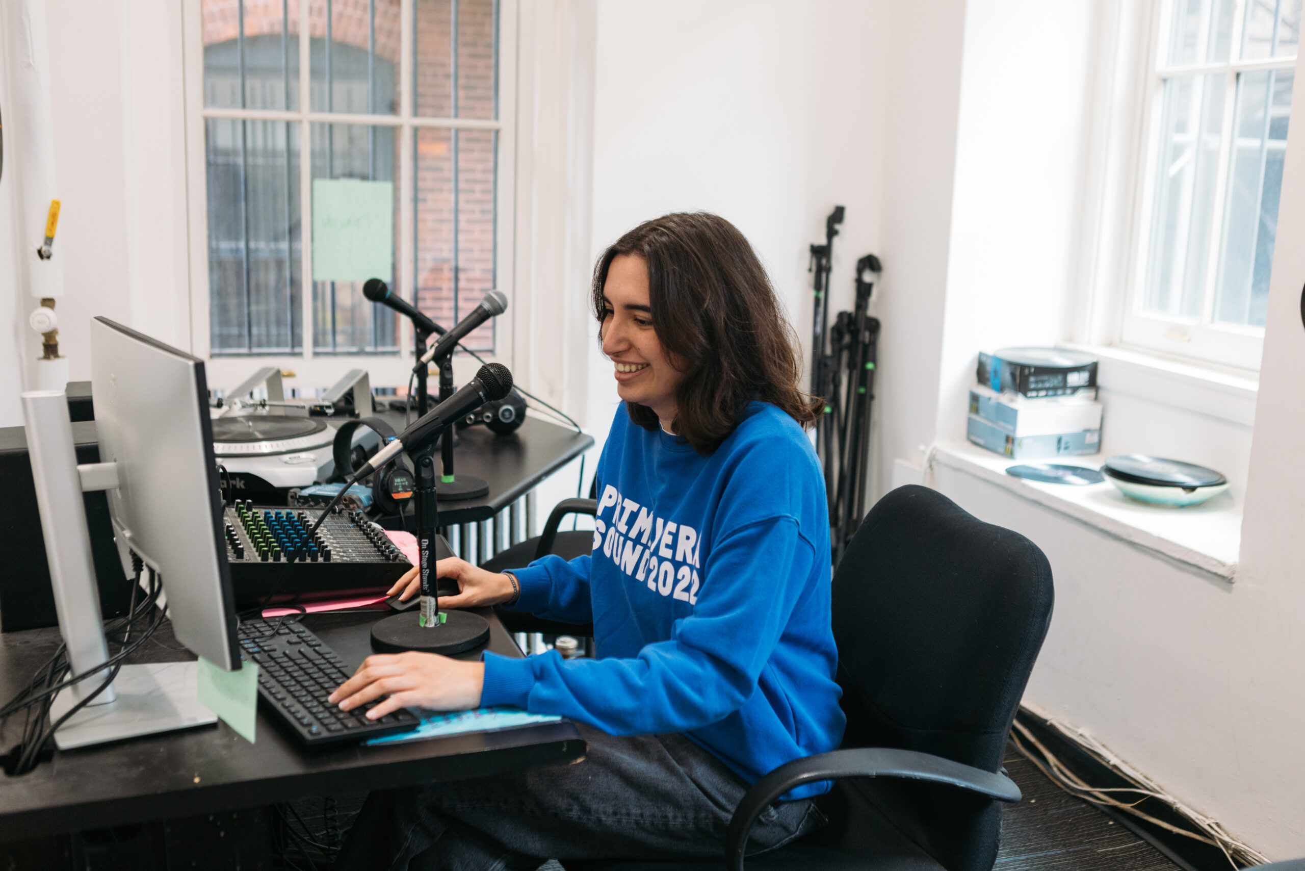A young woman sits at a desk in a recording studio, smiling while typing on a keyboard. She wears a blue sweatshirt with white text. Two microphones are positioned on the desk, along with a sound mixing device and additional equipment in the background. Natural light enters through a window, illuminating the space.
