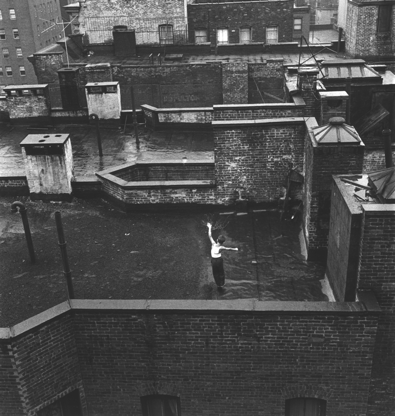 In a black and white photo, a young boy on a Brooklyn rooftop walks as though balancing on a balance beam.