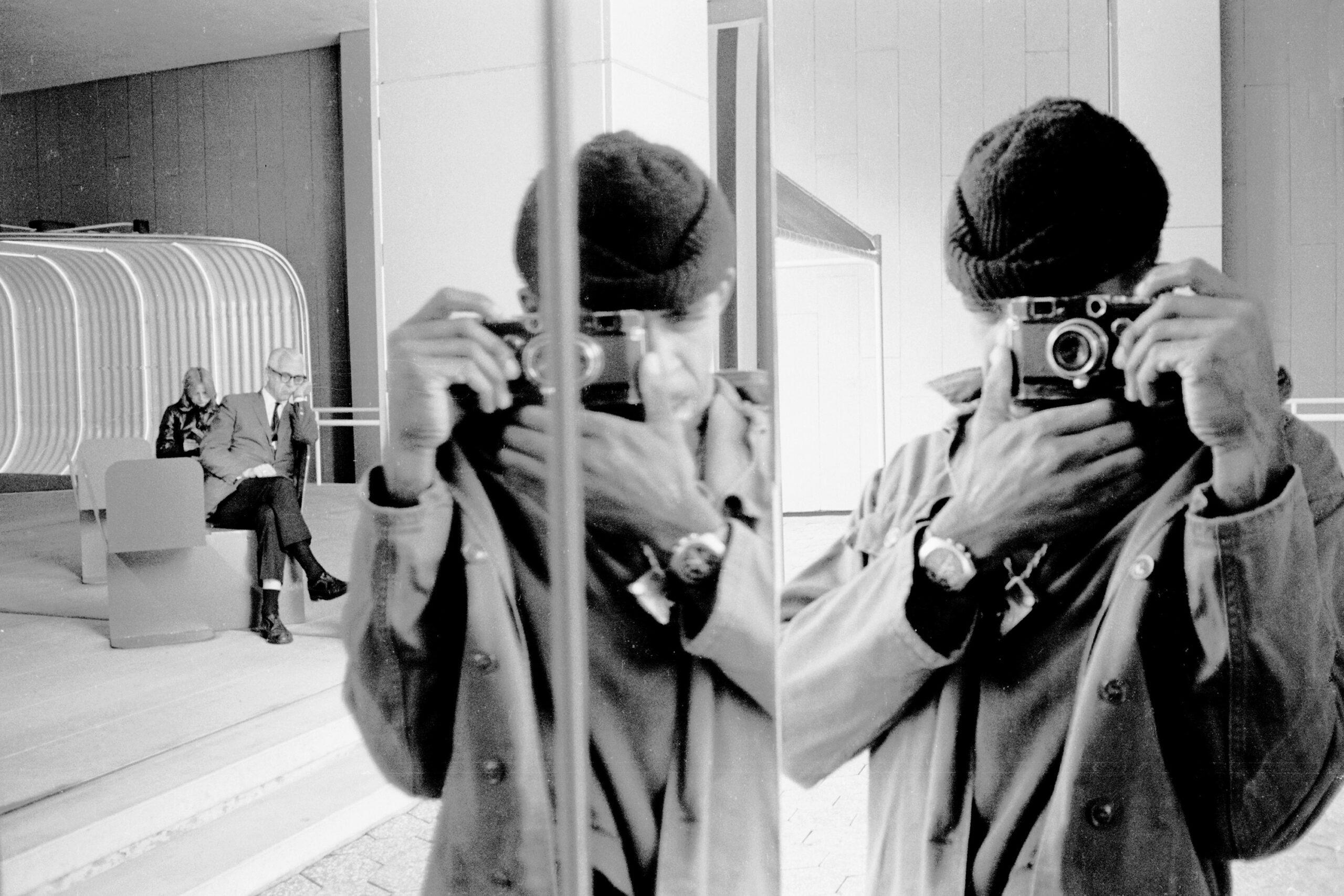 A black and white photograph capturing a person taking a selfie with a vintage camera. The reflection shows their hand holding the camera and another figure seated in the background, appearing deep in thought.