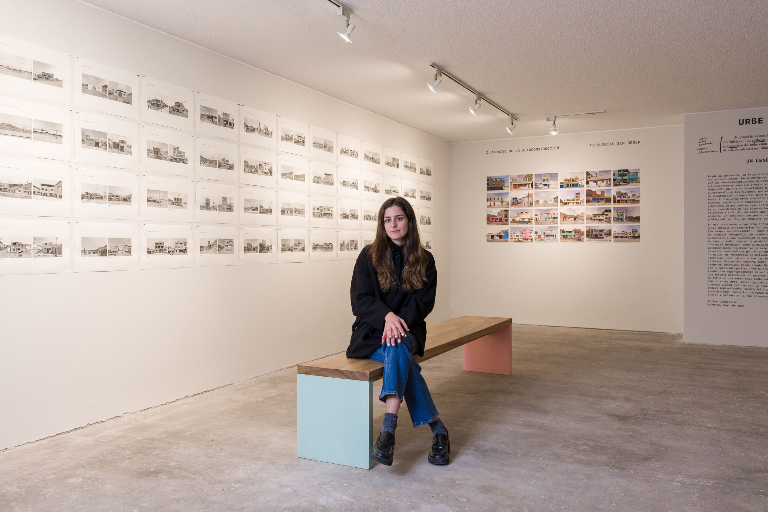 Rafaella Castagnola sits on a bench in an art gallery, wearing a black coat and jeans. Behind her, multiple black-and-white architectural drawings are displayed on the wall. To her right, a collection of colorful images is also mounted, and there is text on the wall related to the exhibition.
