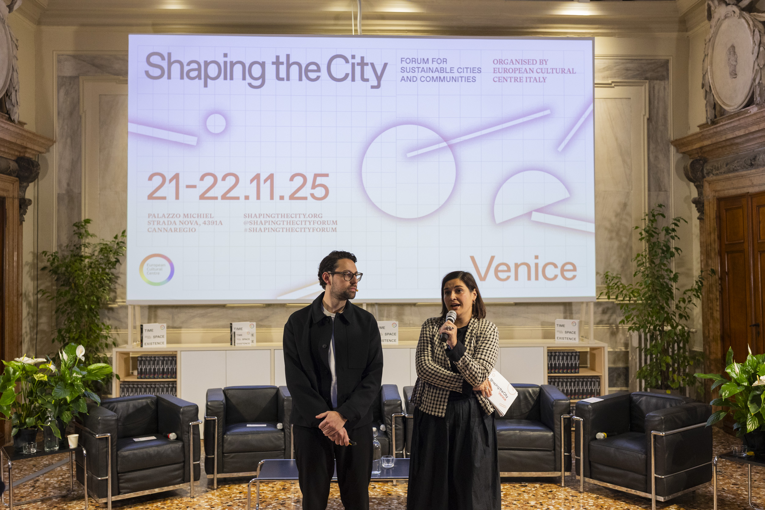 A woman holding a microphone speaks while standing next to a man in glasses at an event. Behind them, a large screen displays "Shaping the City" alongside event details including dates (21-22.11.25), location (Palazzo Michiel, Venice), and hashtags. The setting features chairs, plants, and shelves filled with books.
