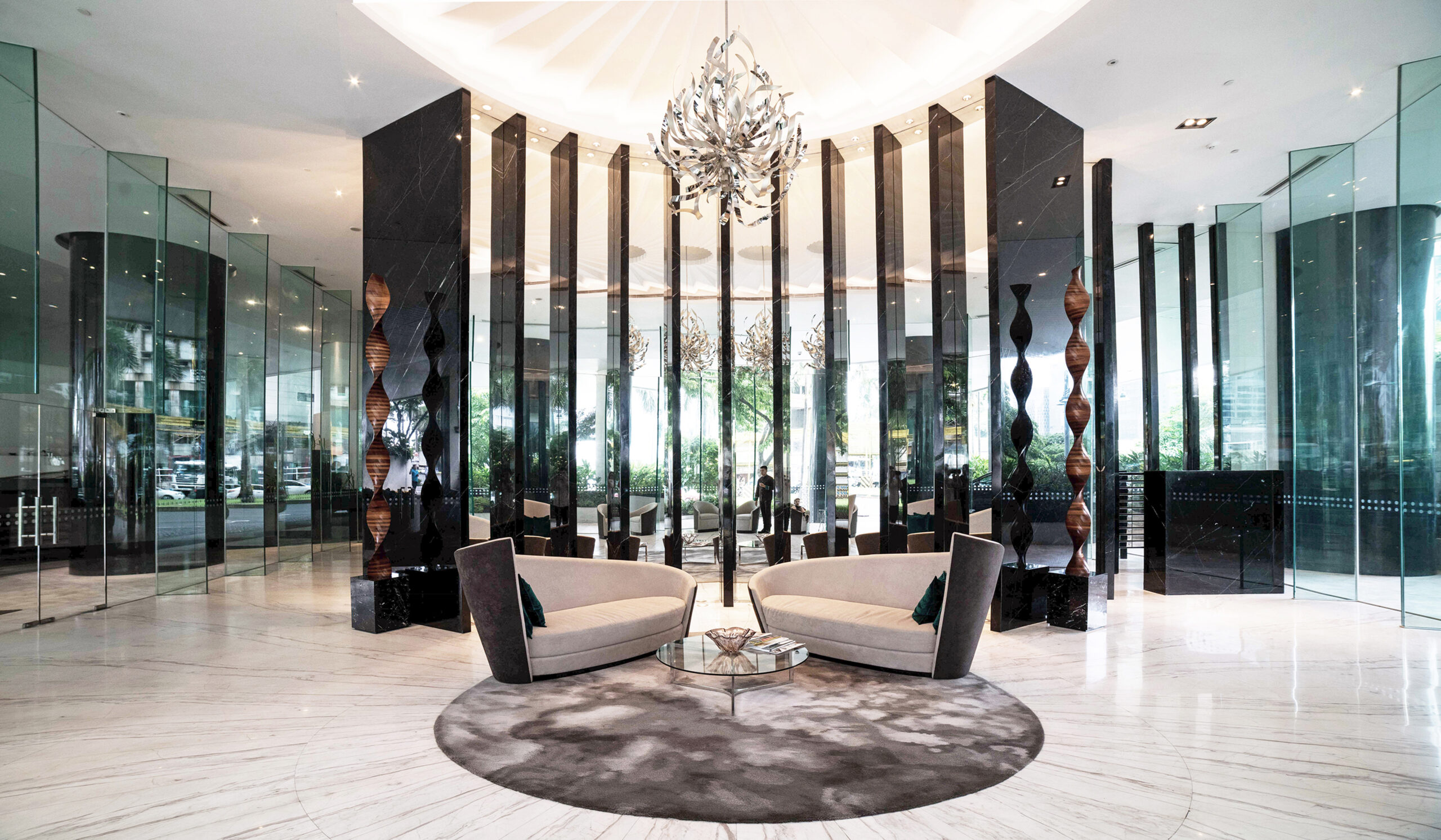 A modern hotel lobby featuring two curved sofas with teal cushions, positioned around a glass coffee table on a circular gray rug. The space is adorned with sleek black columns and large glass panels, reflecting natural light and the greenery outside. A striking chandelier hangs above, enhancing the spacious and elegant atmosphere.