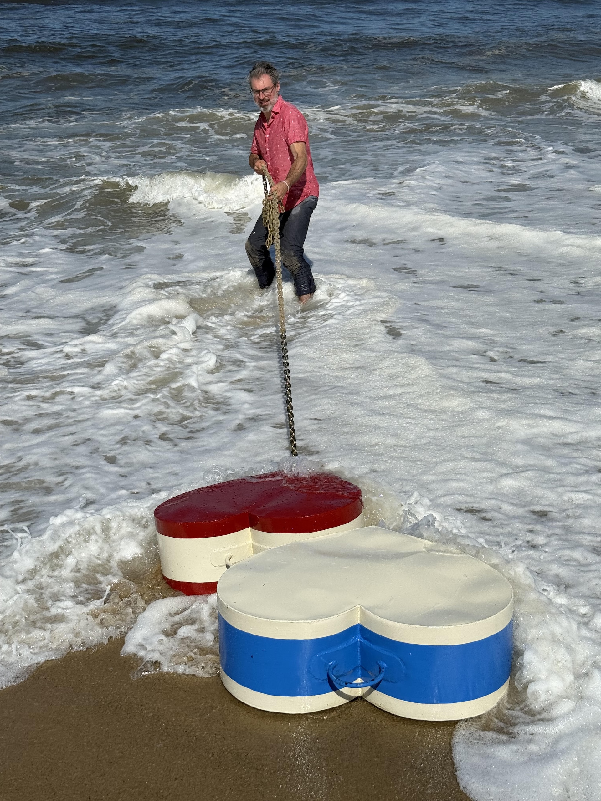 A man in a pink shirt stands in shallow ocean waters, pulling on a chain connected to two large, heart-shaped objects on the beach. One object is red and white, while the other is white and blue. Waves wash over the beach, creating foam around the objects and the man.