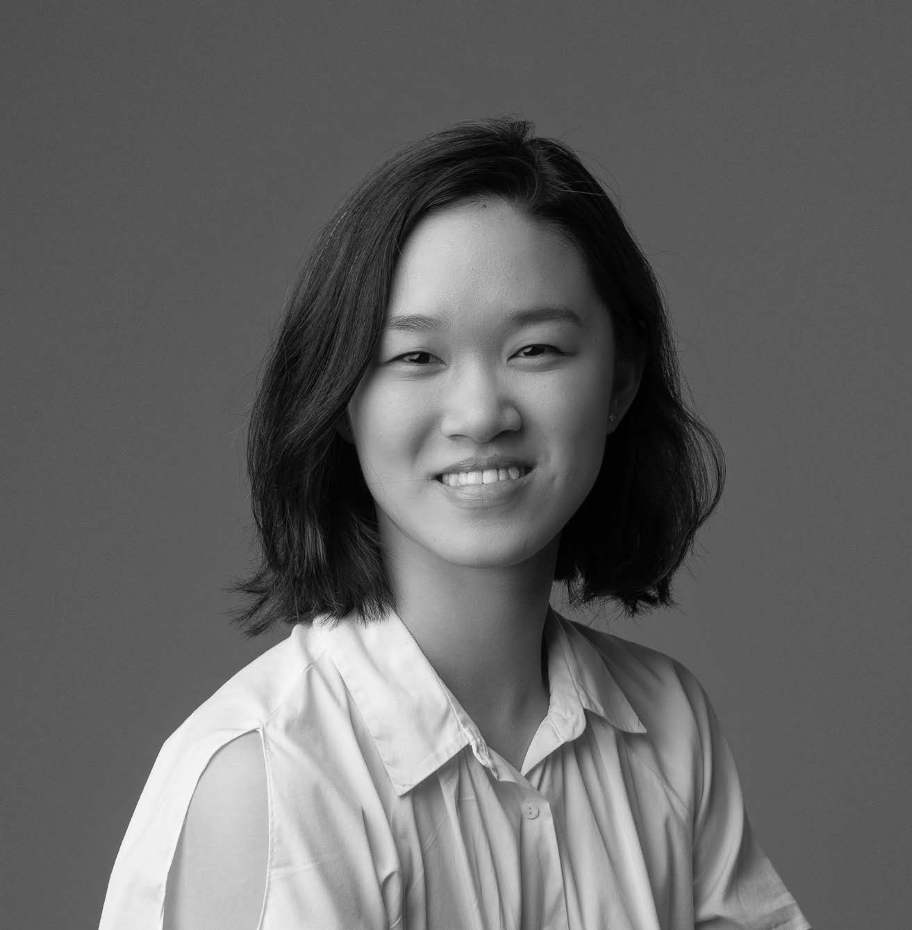 A smiling young woman with shoulder-length dark hair is facing the camera. She is wearing a white collared shirt. The background is a solid gray color, creating a simple and clean portrait.