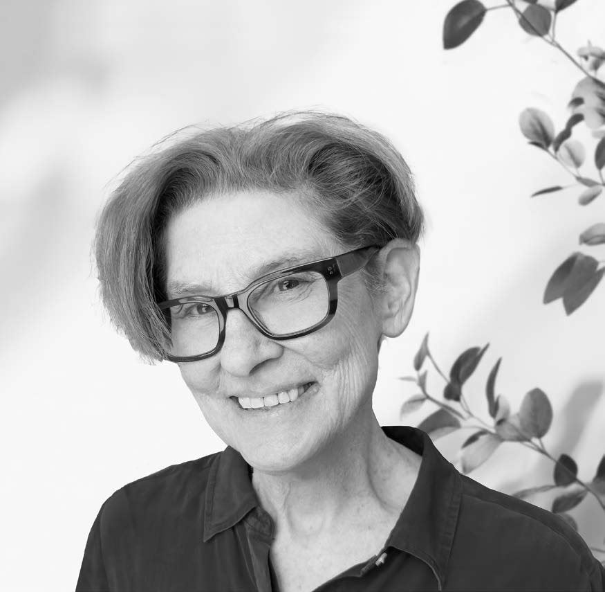 A smiling older woman with short, styled gray hair and large black glasses is facing the camera. She is wearing a dark collared shirt. The background is light and features soft shadows, with green foliage visible on the right side.