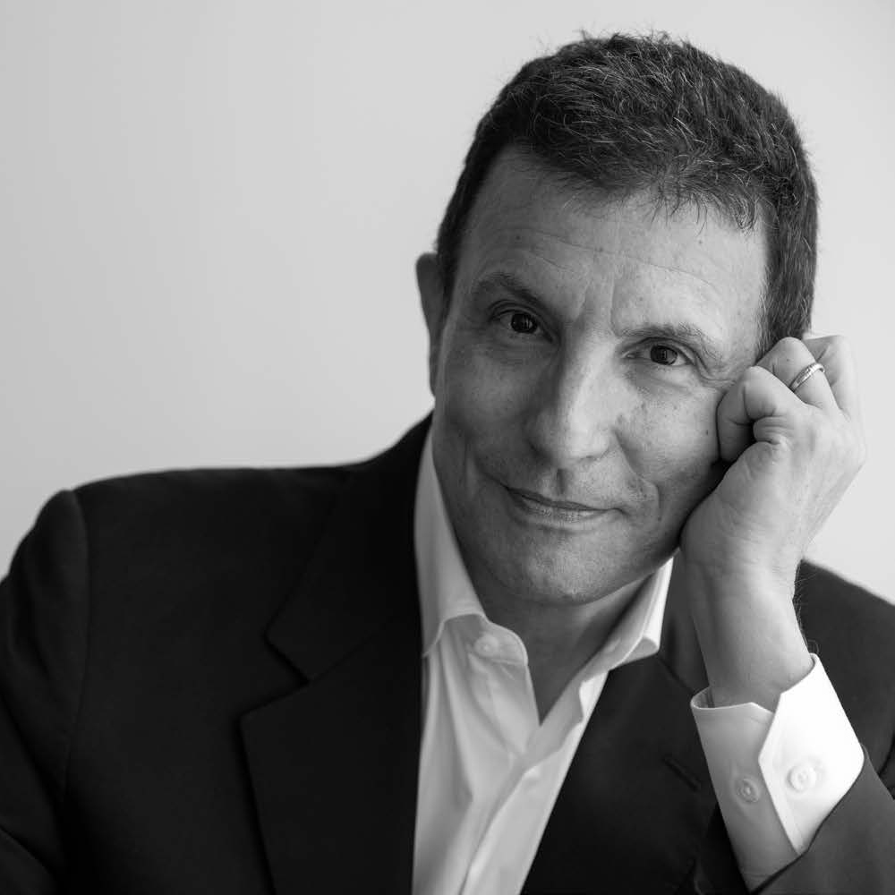 A man in a black suit and white shirt rests his chin on his hand, smiling softly. The photo is in black and white, highlighting his features. He has short, dark hair and appears engaged and thoughtful. The background is a light, soft gradient.