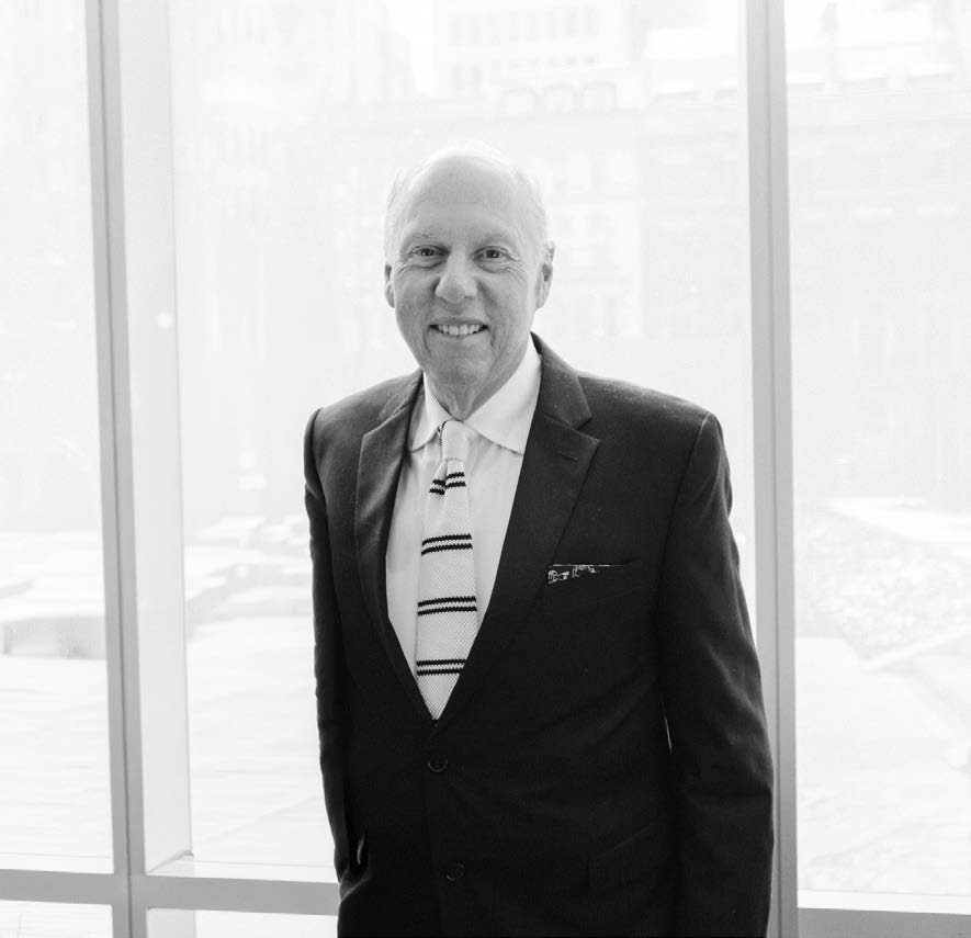 A smiling man in a formal black suit with a white shirt and a striped tie stands by a large window. The background is softly blurred, suggesting an outdoor view. The man has short, light-colored hair and is facing the camera, presenting a friendly demeanor.