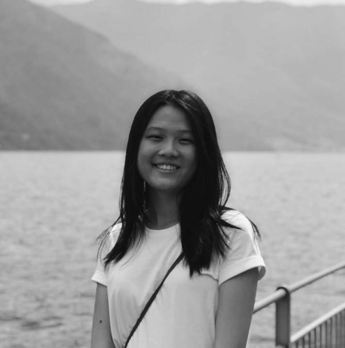 A young woman with long black hair smiles while standing near a body of water. She is wearing a white shirt and has a strap across her shoulder. In the background, blurred mountains rise above the water, creating a serene landscape in black and white.