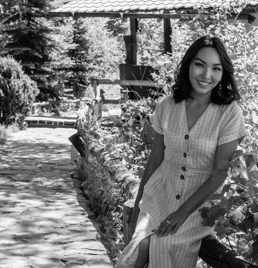 A woman sits gracefully on a wooden fence, smiling as she poses for the camera. She wears a light-colored, button-up dress with short sleeves, featuring a subtle striped pattern. The background features a pathway made of stone and lush greenery, suggesting a serene outdoor setting. Sunlight filters through the trees, creating a warm atmosphere.