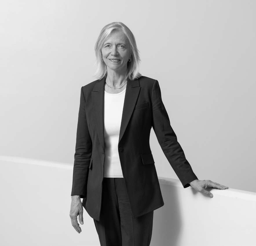 A woman stands confidently, wearing a dark suit over a light-colored shirt. She has shoulder-length blonde hair and is smiling while resting one hand on a white surface. The background is a smooth, light color, giving a clean and professional appearance.