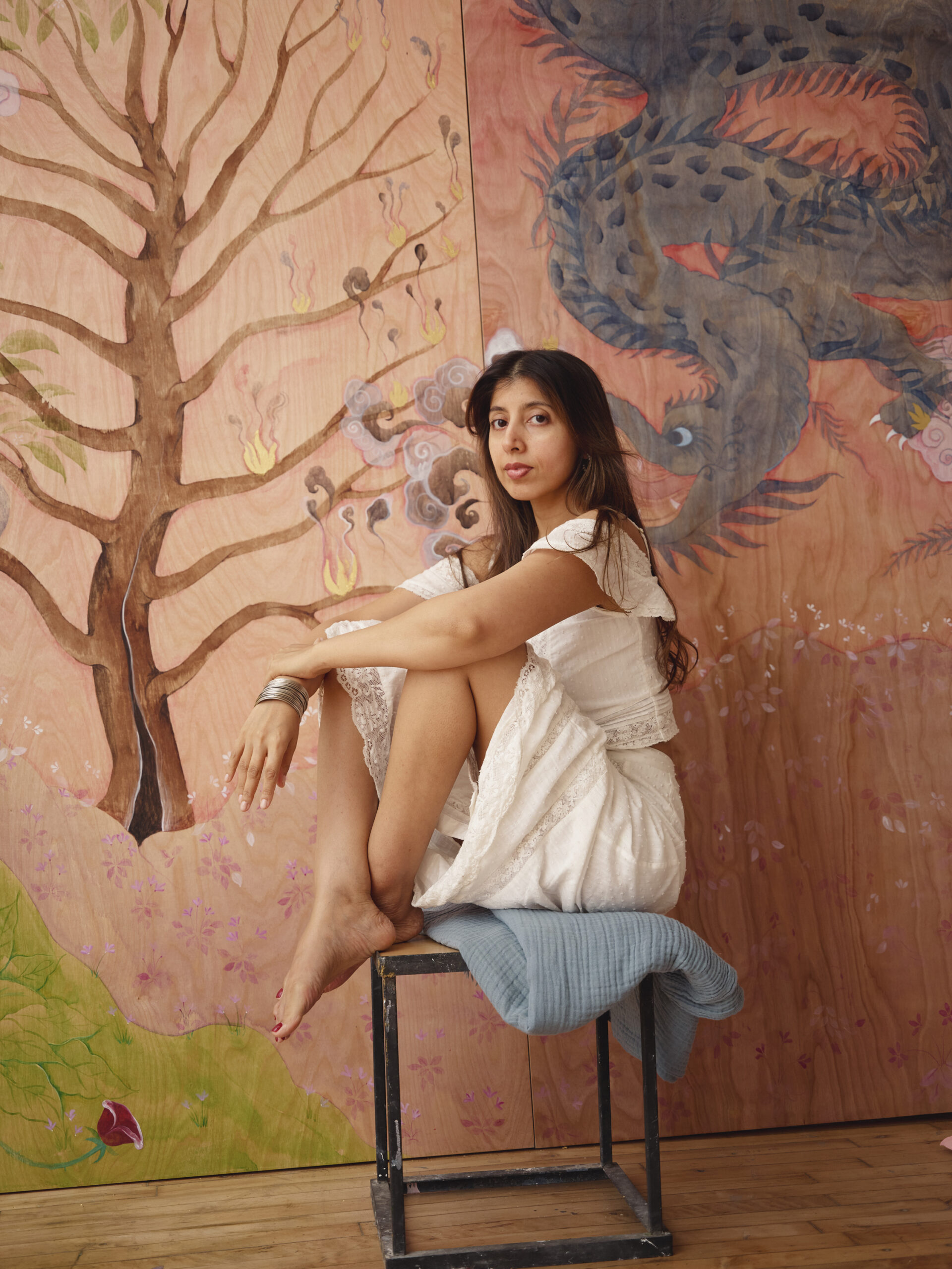 Hiba Schahbaz poses against a colorful mural featuring a tree and a dragon. She wears a white dress and silver bangles, with her bare feet resting on a blue cushion atop a metal stool. The background art includes soft pinks and greens, with floral details and stylized elements.