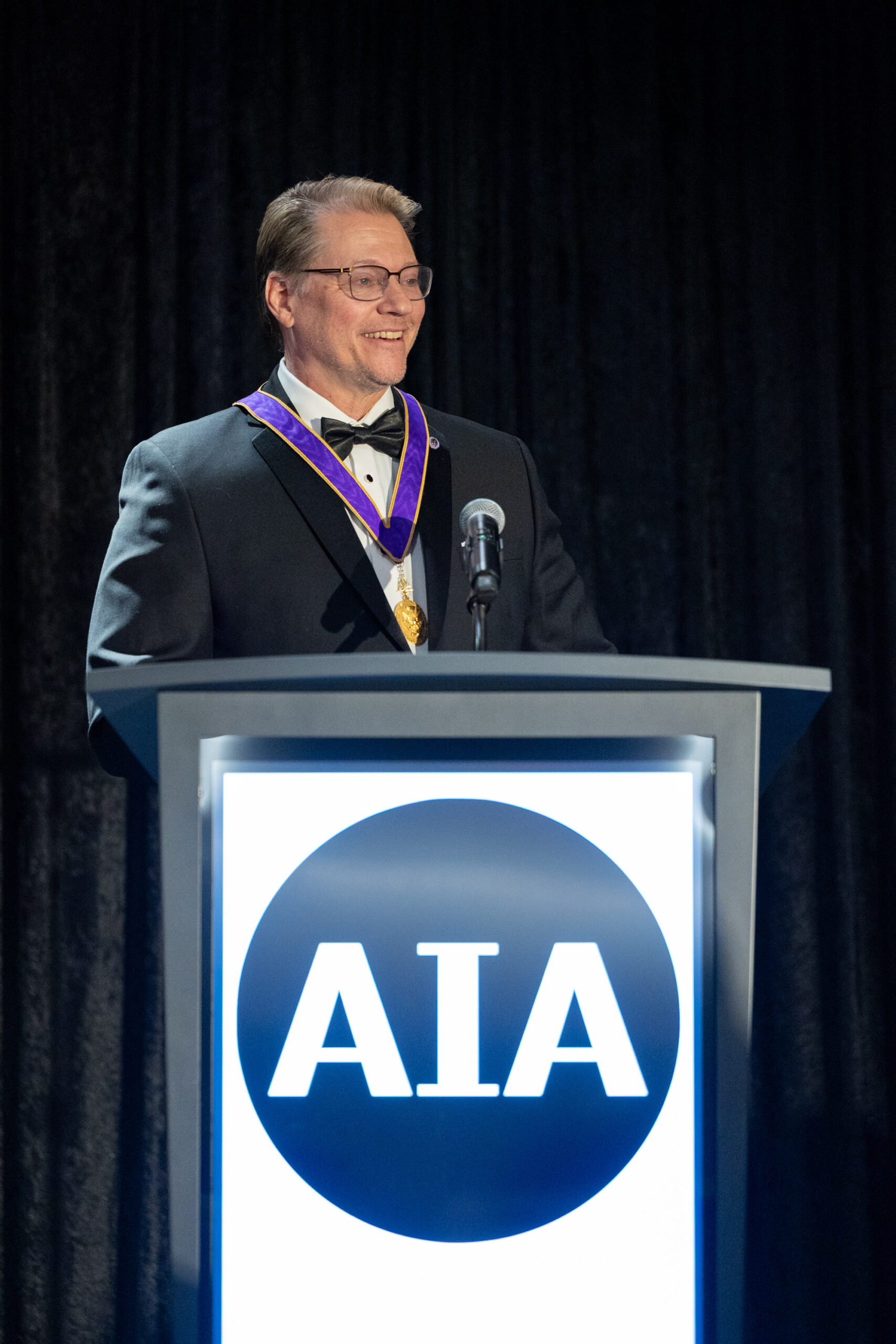 Illya Azaroff stands and smiles at an AIA podium
