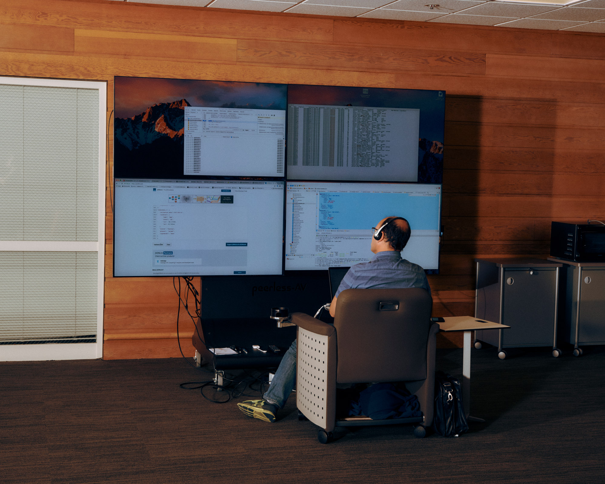 A person sitting in a chair with headphones is facing a setup of four large monitors displaying various computer screens, including code and web pages. The background features wooden walls, and there are storage cabinets to the right of the image. The workspace is modern and organized, with a small table beside the chair.