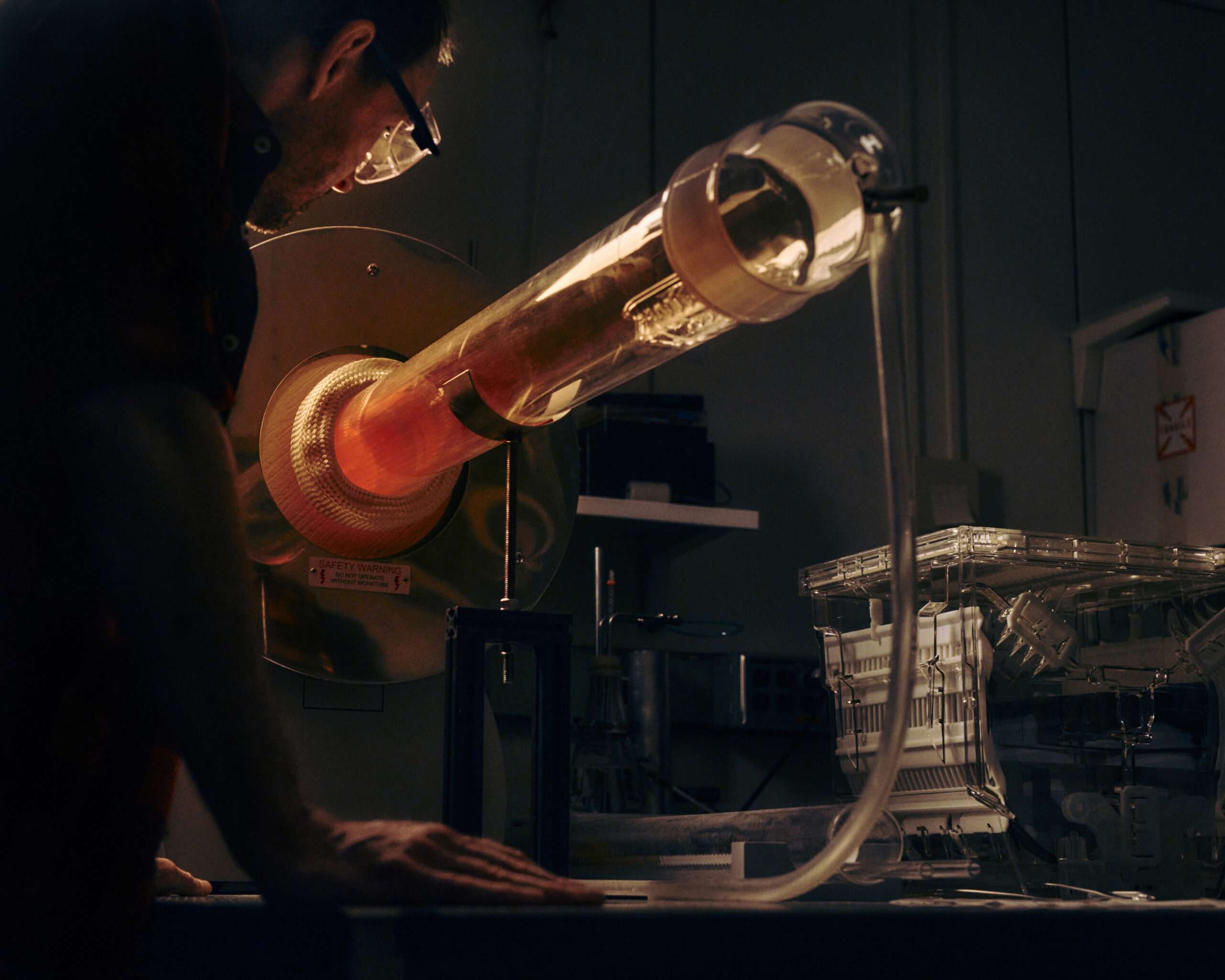 A close-up image of a person leaning over a laboratory table, observing a large glass apparatus that emits a warm orange glow. The apparatus is cylindrical, with a metallic base and a tube extending from it. The setting is dimly lit, emphasizing the glowing section of the device.