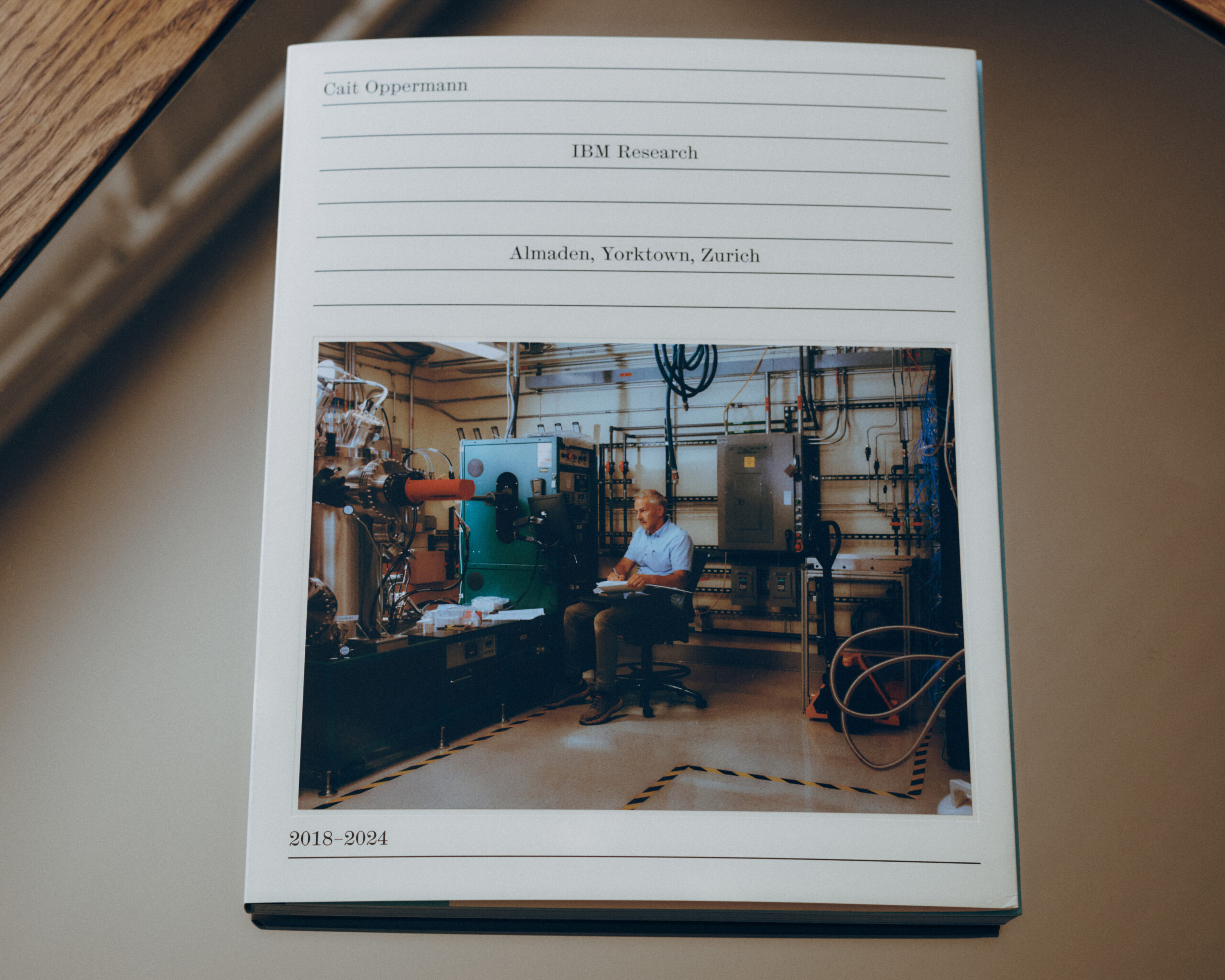 A book cover featuring a photo of a man seated at a table with equipment in a research lab. The background displays various machines and scientific instruments. The book's title at the top reads "Cait Oppermann," with "IBM Research" below it, and mentions "Almaden, Yorktown, Zurich." The year range "2018–2024" is noted at the bottom.